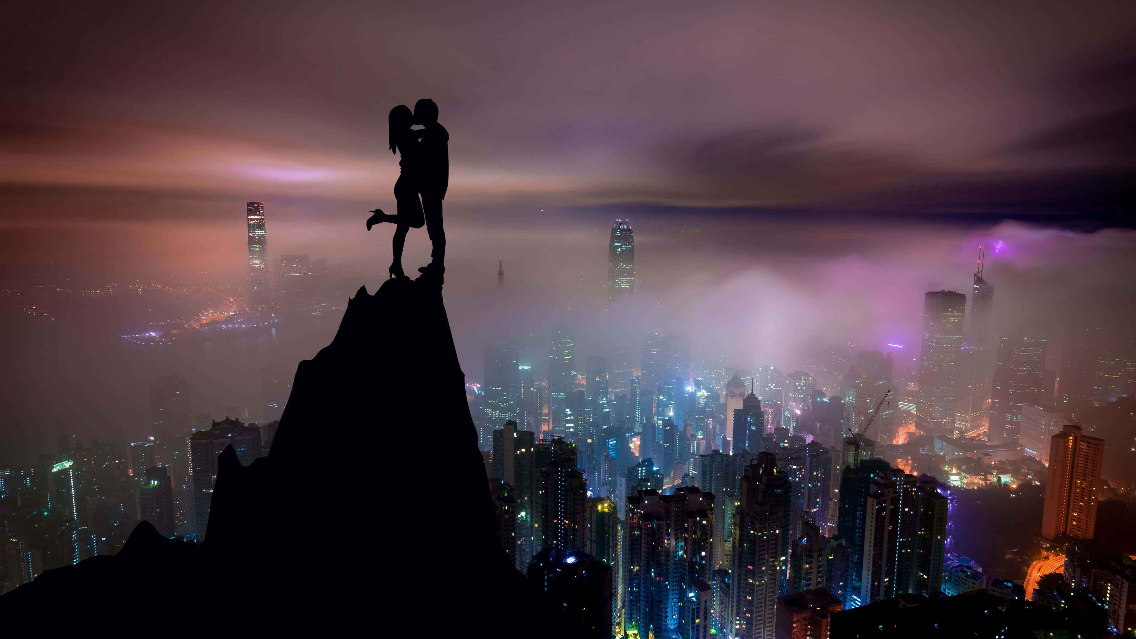4K Love 桌布 壁紙 — 一對剪影情侶在懸崖頂上擁抱，俯瞰暮色中霧氣瀰漫、霓虹閃爍的城市景觀。