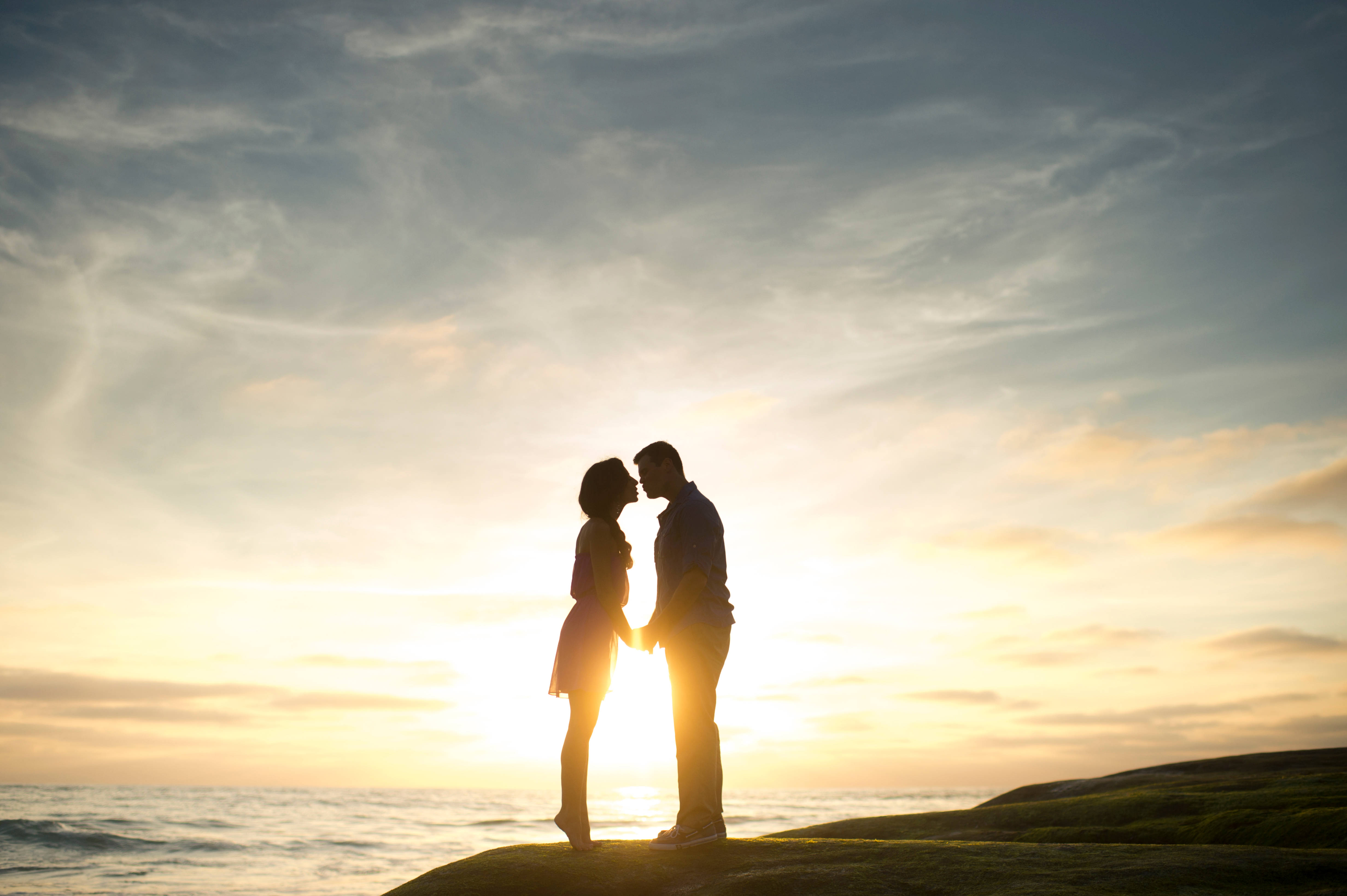 Romantic Couples 桌布 壁紙 — 一對剪影情侶在日落時分站在懸崖邊，手牽手，在發光的天空和海洋背景下傾身親吻。