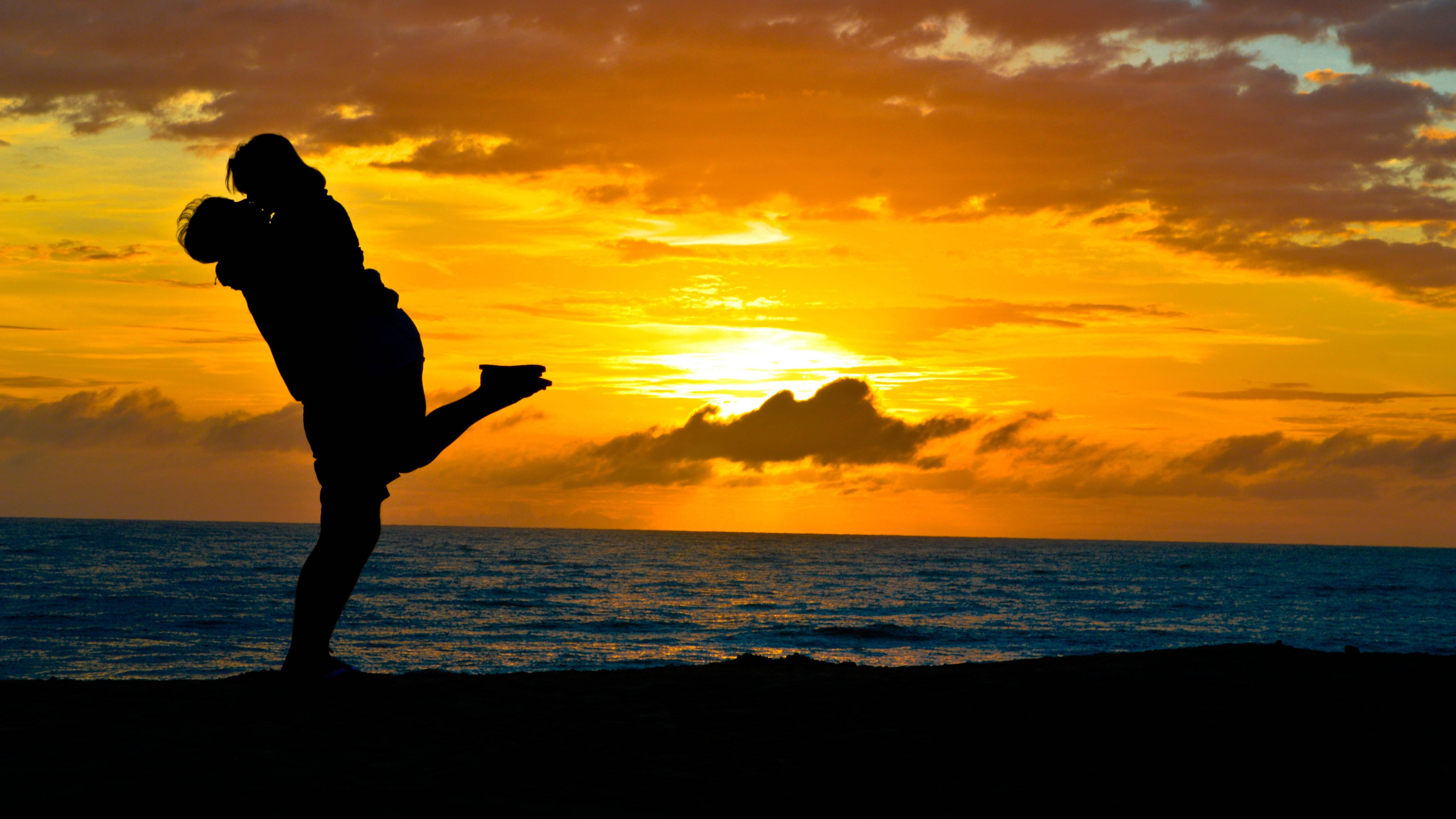 Silhouette 桌布 壁紙 — 一對剪影情侶在海灘上擁抱，夕陽以火熱的橙色和黃色色調沉入海洋。