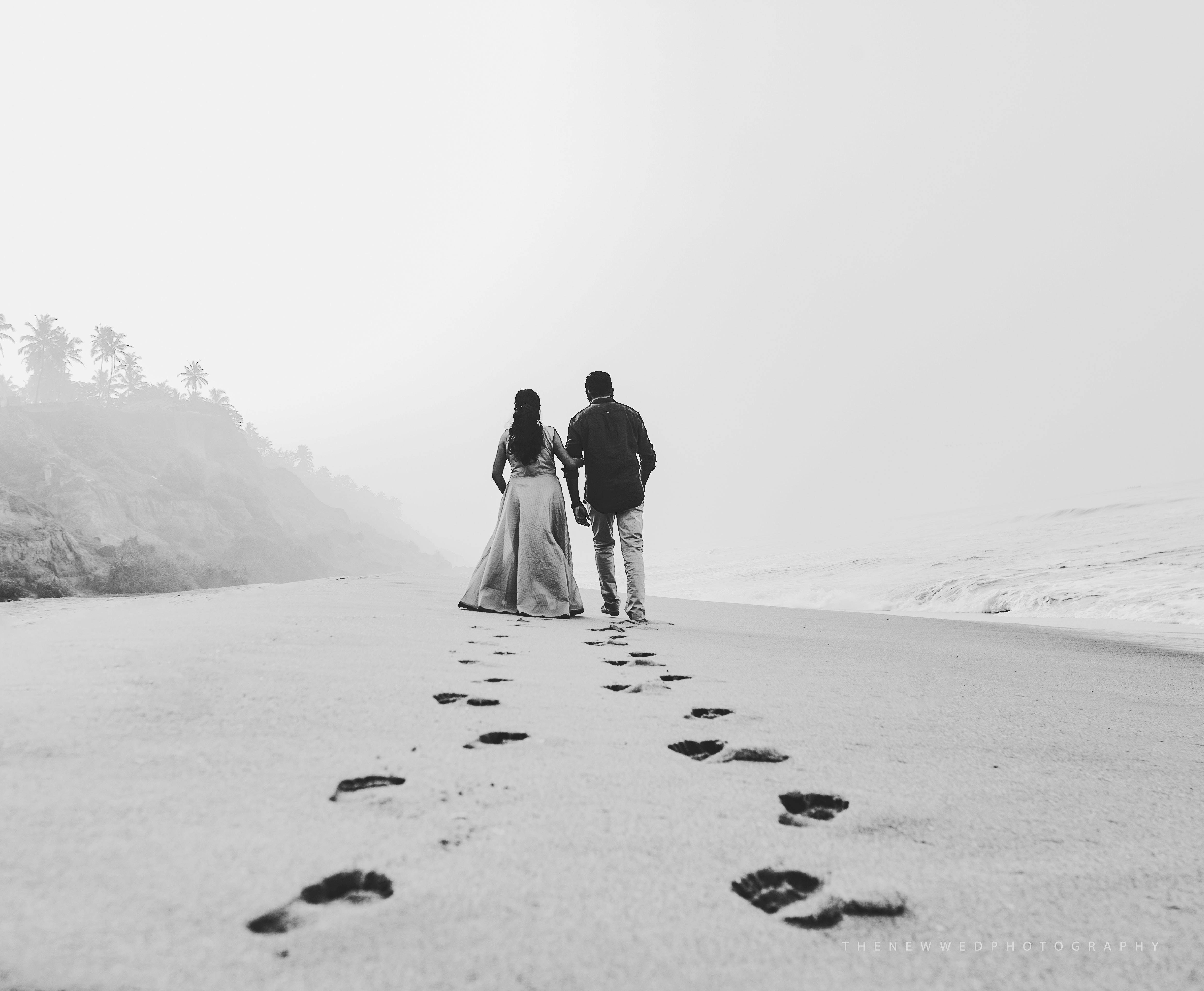 Couples 桌布 壁紙 — 一對單色調的情侶手牽手沿著霧濛濛的海灘漫步，在沙灘上留下腳印，遠處的山丘上有棕櫚樹。