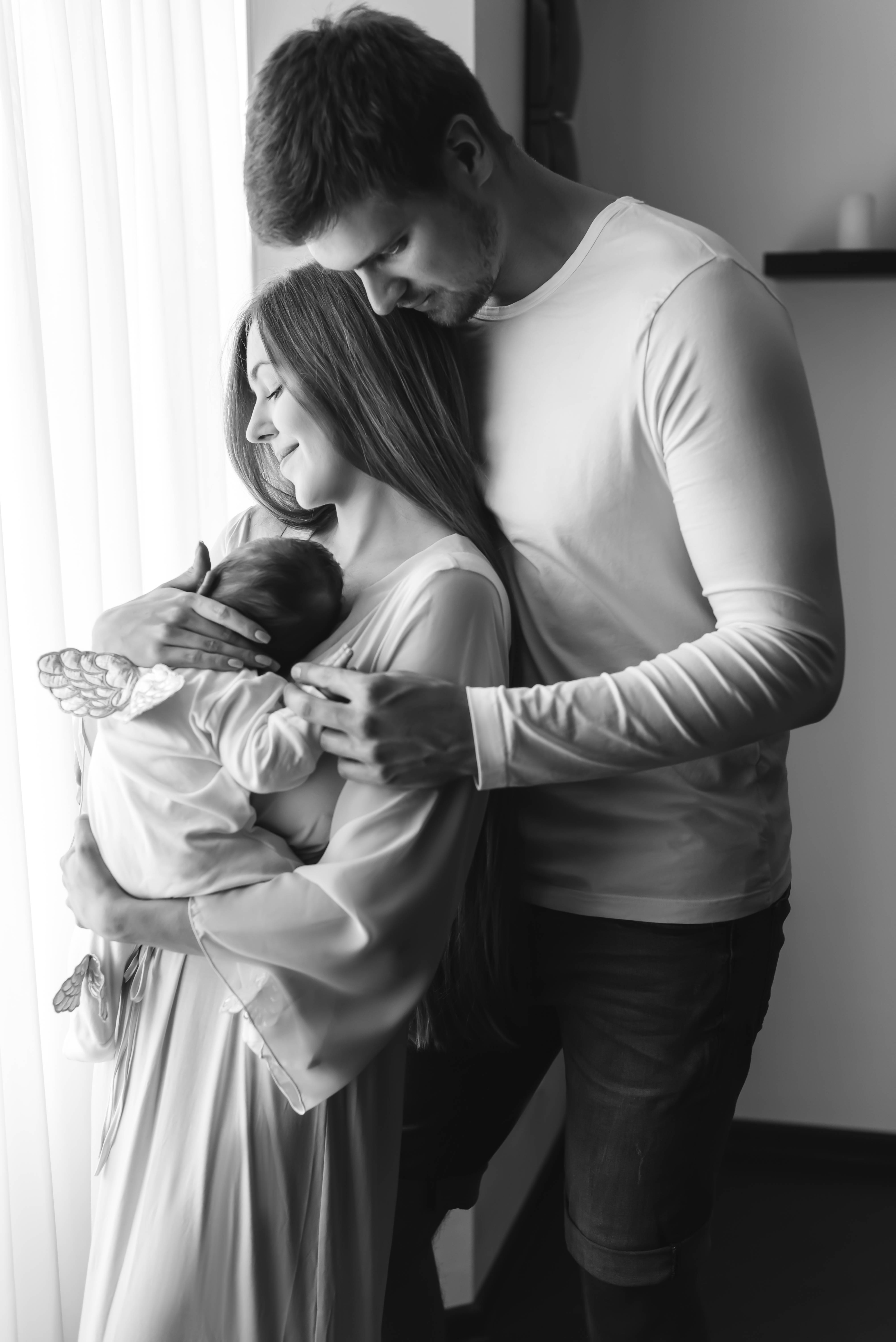 Family 桌布 壁紙 — 一對夫婦在窗邊擁抱新生嬰兒的溫柔黑白照片。