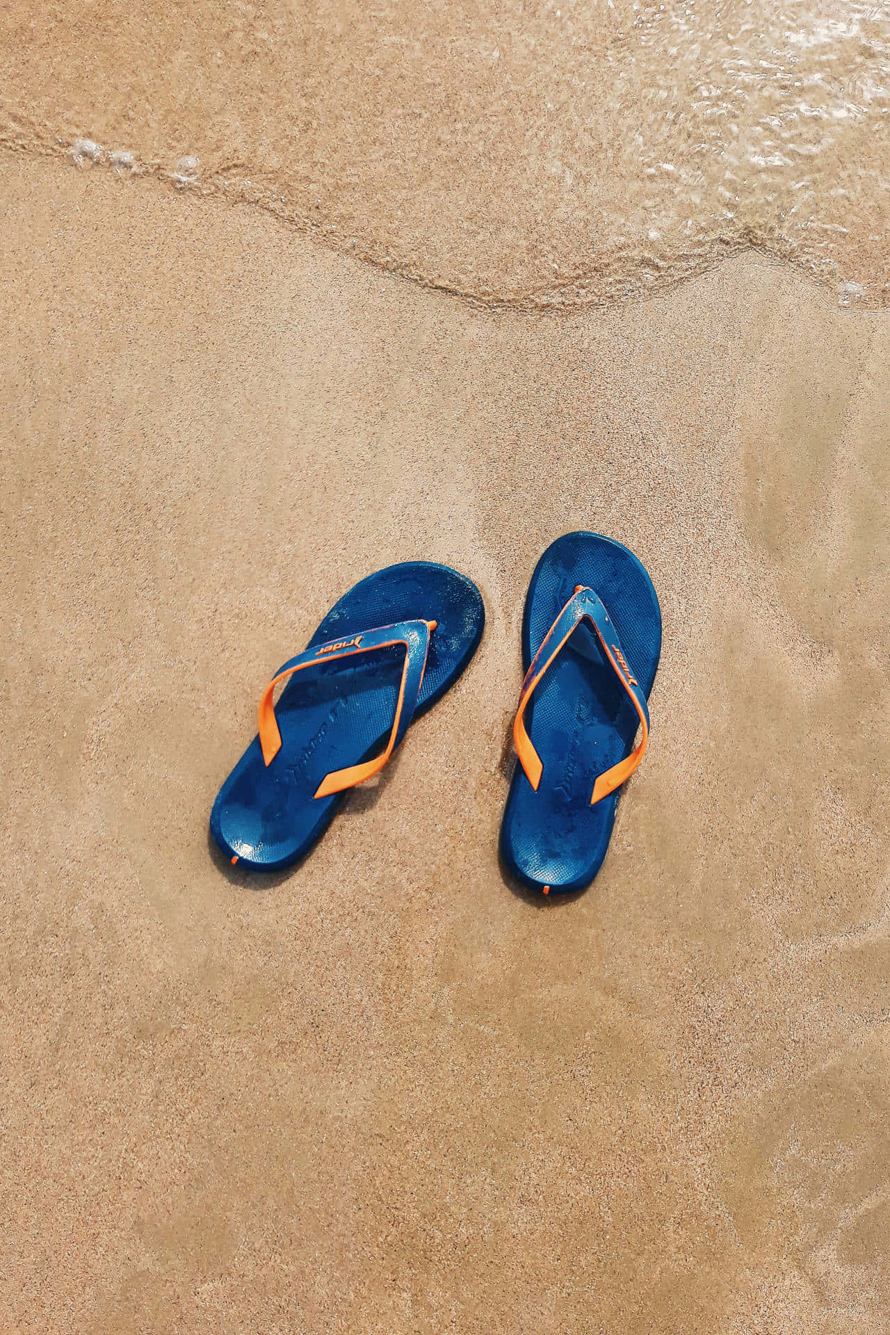Beach Sandals 桌布 壁紙 — 一對帶有橙色帶子的藍色夾腳拖放在濕沙上，溫柔的海浪在附近拍打。