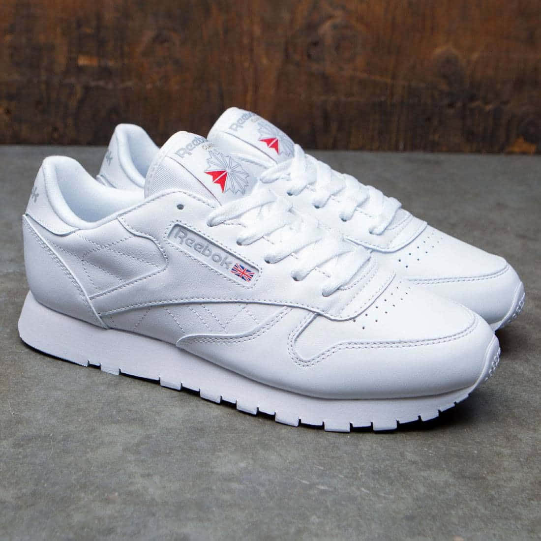 Reebok 桌布 壁紙 — 一對帶有紅色和藍色點綴的純白色Reebok運動鞋，放在灰色混凝土地板上，背景是質樸的木頭。