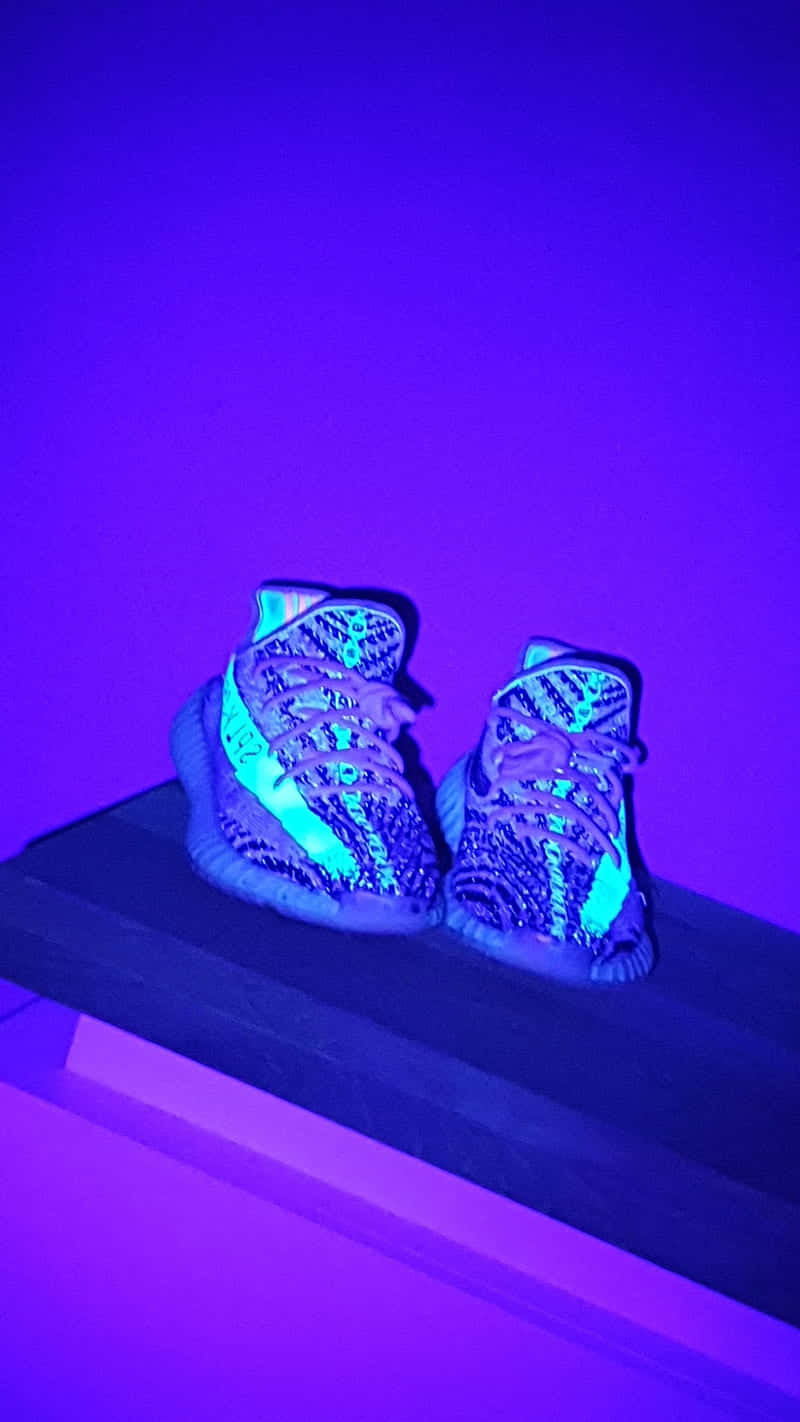 Yeezy 桌布 壁紙 — 一對帶有螢光藍色點綴的發光運動鞋放在深色木架上，背景是充滿活力的紫色牆壁。