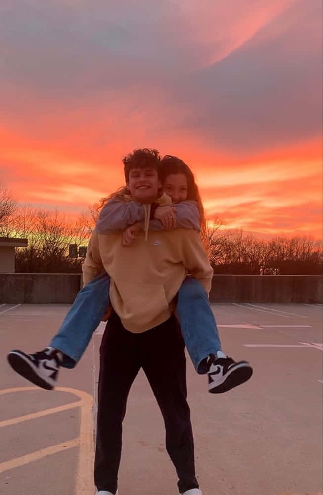 Girlfriend And Boyfriend 桌布 壁紙 — 一對年輕情侶在 vibrant 的日落天空下，男方揹著女方，共享歡樂時光。