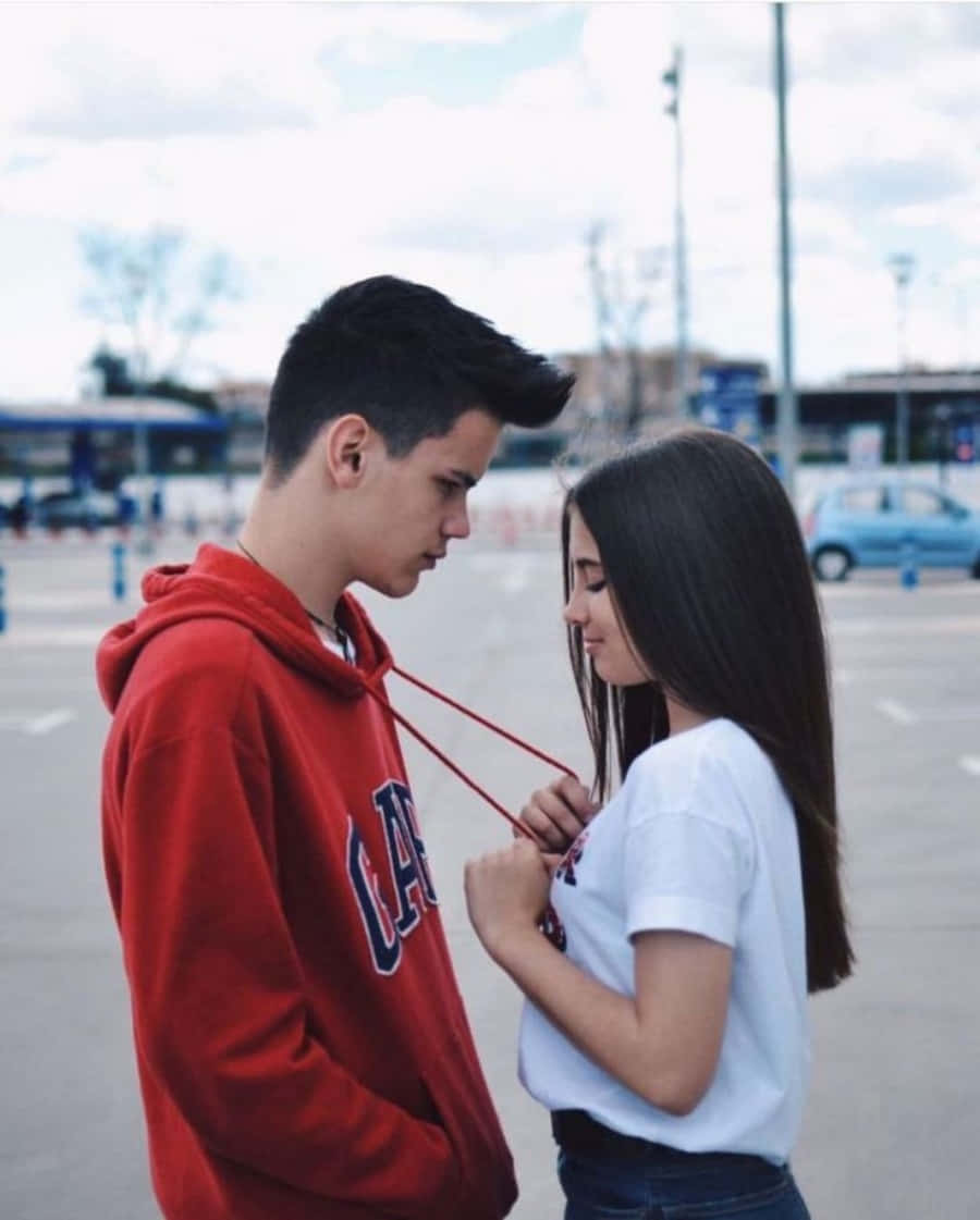 Boyfriend And Girlfriend 桌布 壁紙 — 一對年輕情侶在停車場分享親密時刻，女孩輕輕地將紅色連帽衫的繩子繞在他的脖子上。