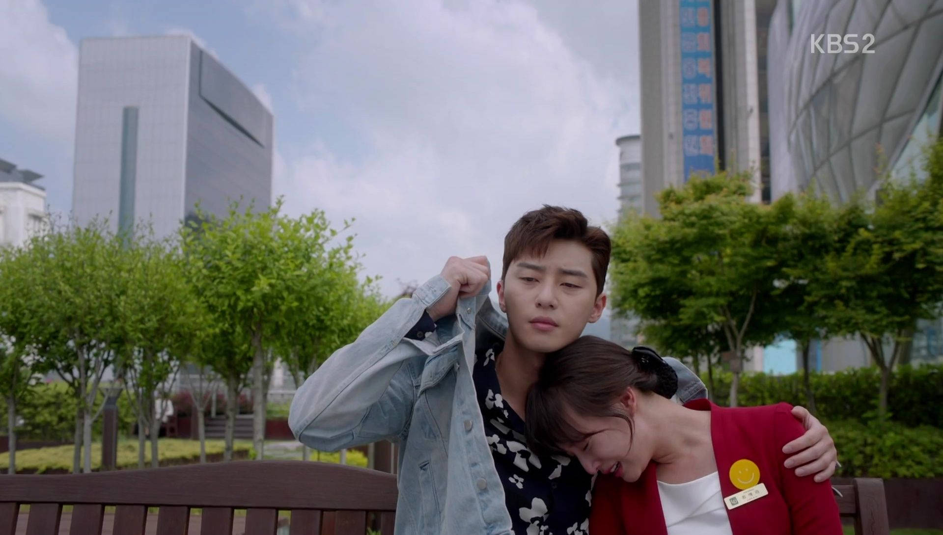 drama 桌布 壁紙 — 一對年輕情侶在公園長椅上分享親密時刻，背景有現代建築和綠樹。