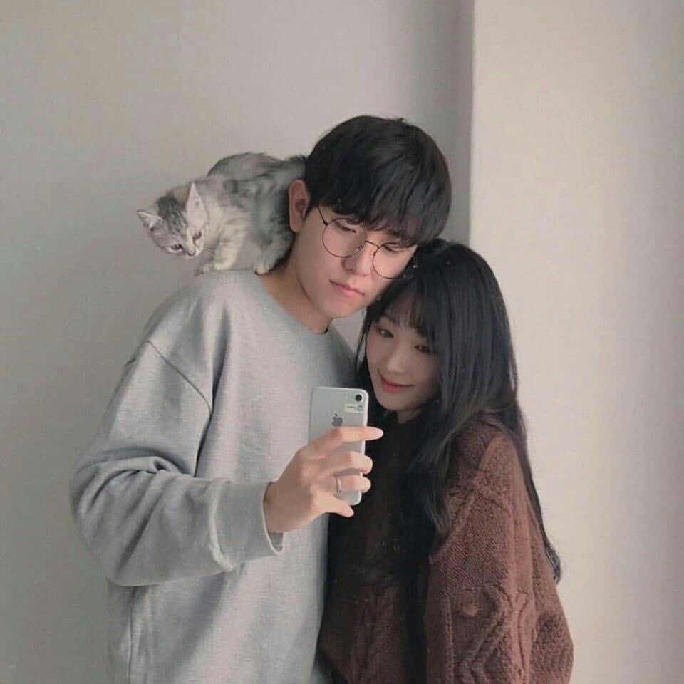 Couples 桌布 壁紙 — 一對年輕情侶在室內自拍，一隻灰色貓咪棲息在男士的肩膀上，背景是素牆。