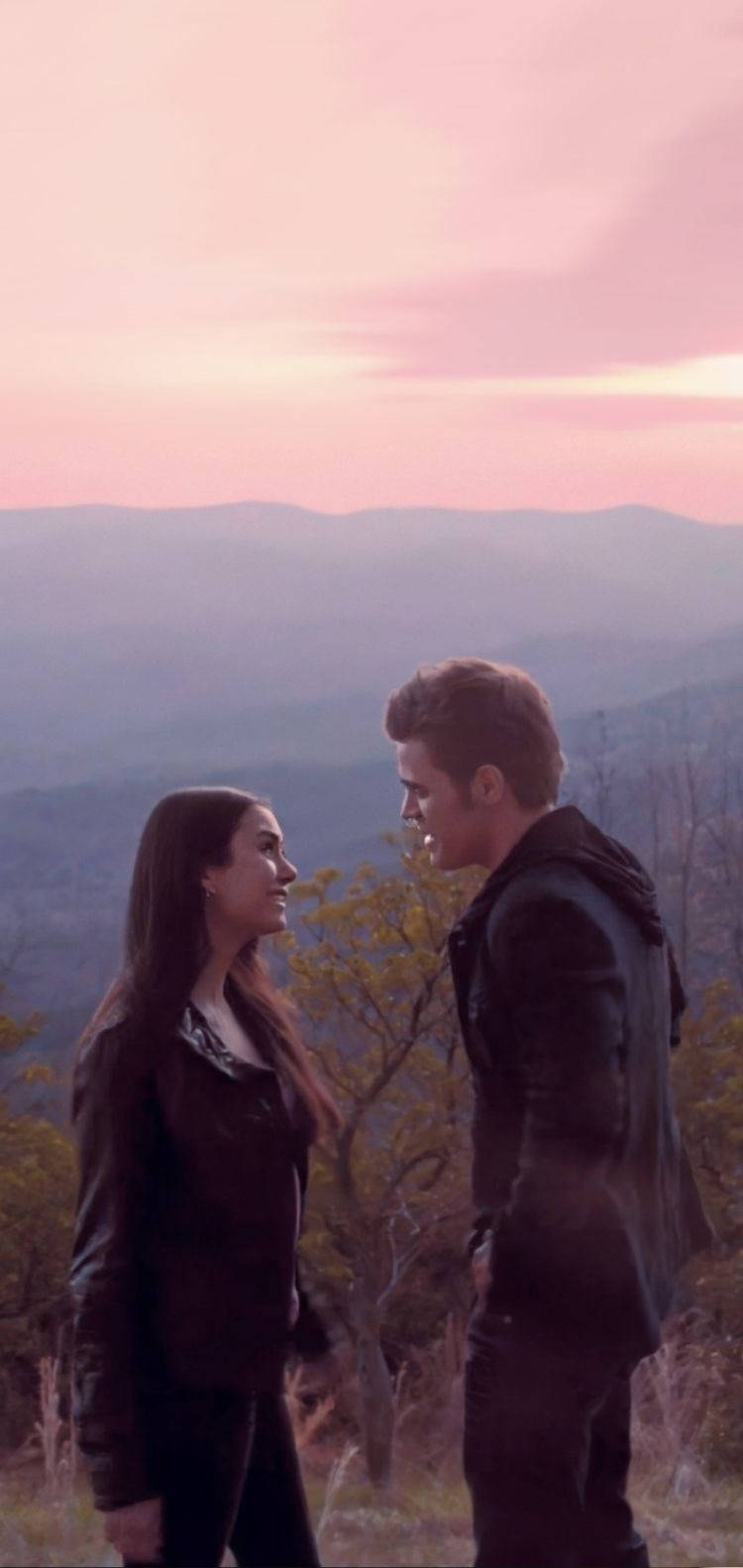 Vampire Diaries 桌布 壁紙 — 一對年輕情侶在日落時分於山坡上面對面站著，在柔和的粉紅色和紫色山脈背景下凝視對方的眼睛。