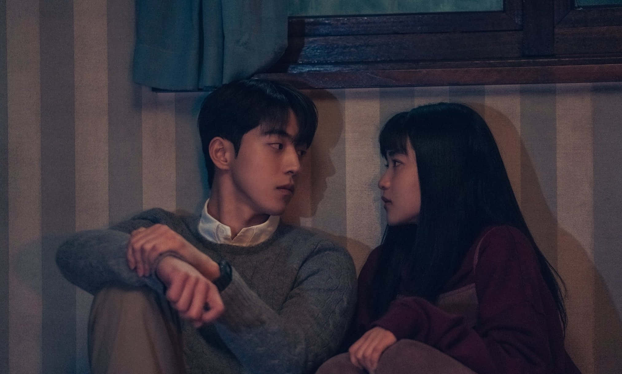 Sad Couple 桌布 壁紙 — 一對年輕情侶在燈光昏暗的房間裡，緊貼著坐在條紋沙發上，分享親密安靜的時刻。