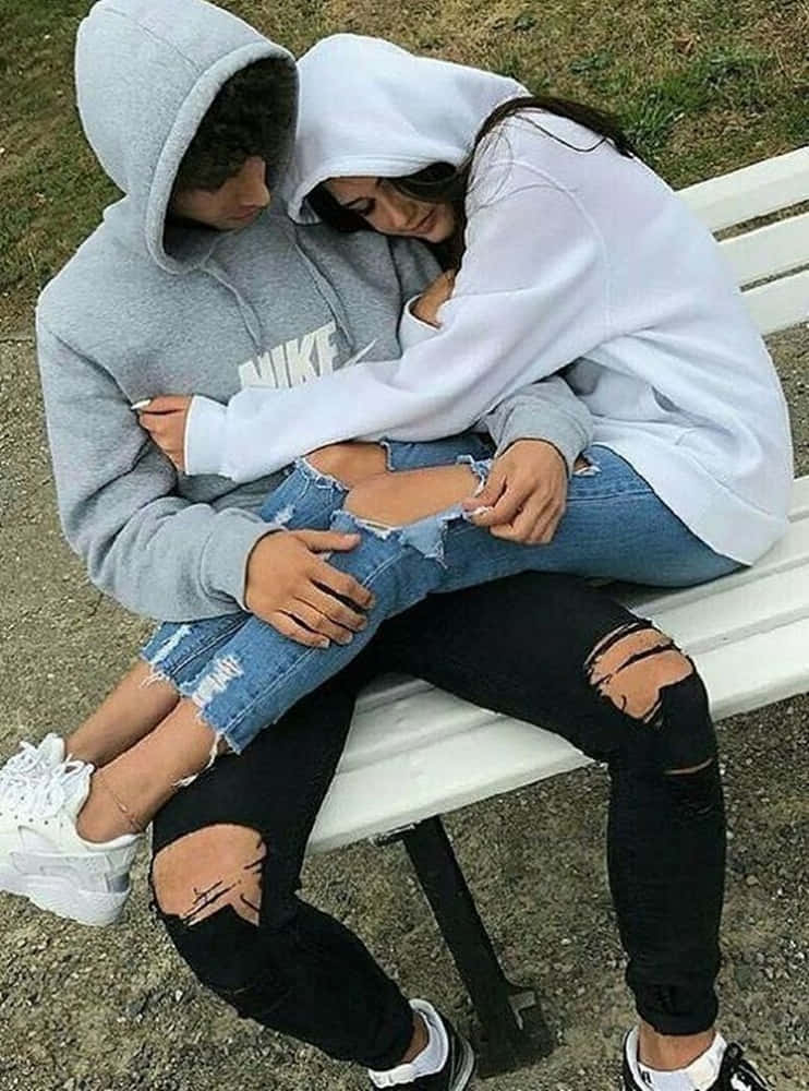 Girlfriend And Boyfriend 桌布 壁紙 — 一對年輕情侶在白色長椅上親密擁抱，穿著連帽衫和破洞牛仔褲，在寧靜的戶外環境中。