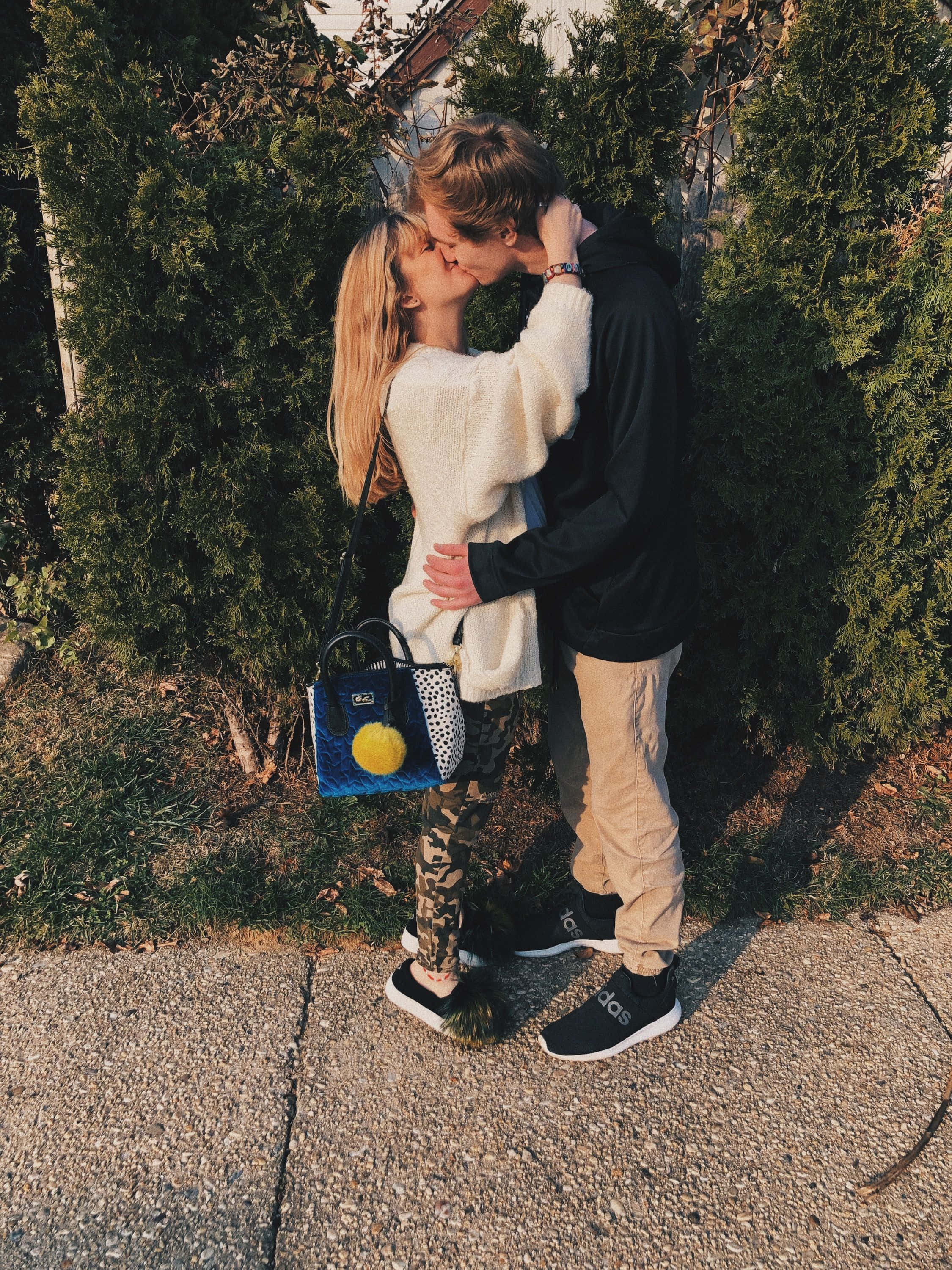 Cute Boyfriend 桌布 壁紙 — 一對年輕情侶在陽光明媚的人行道上，被茂密的綠色樹籬環繞，分享著溫柔的親吻。
