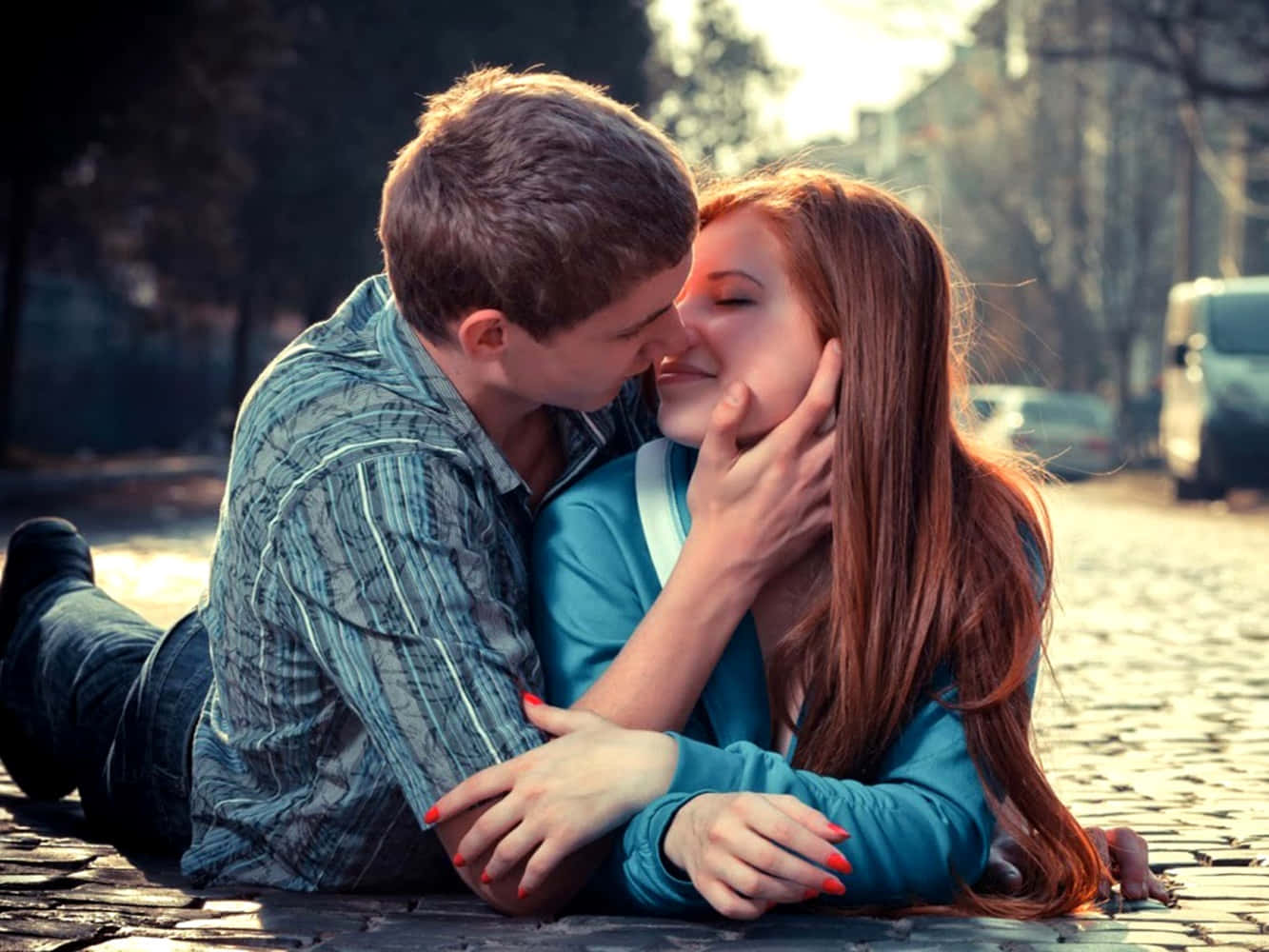 couple love 桌布 壁紙 — 一對年輕情侶在陽光明媚的鵝卵石街道上親密接吻，背景是模糊的汽車和樹木。