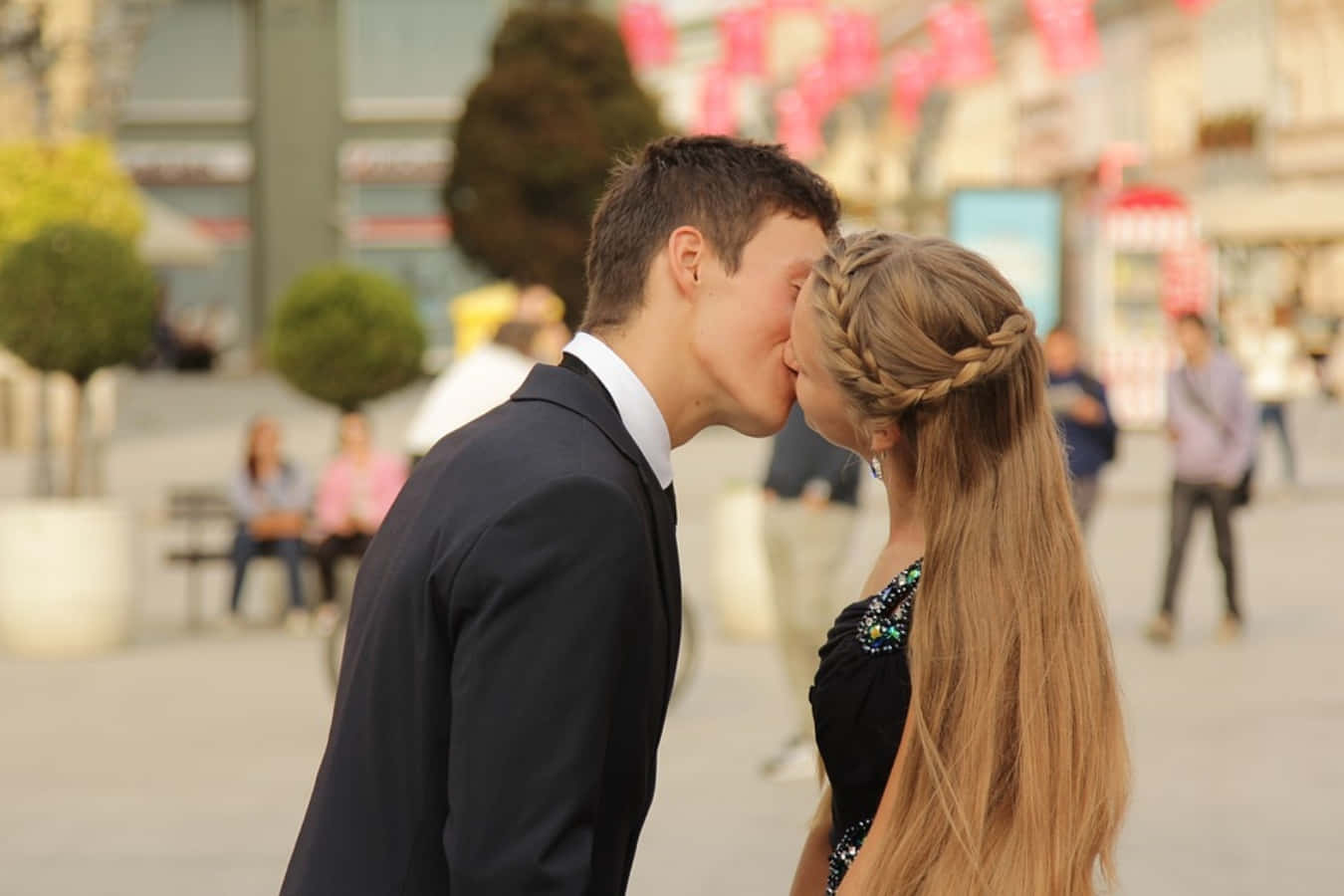 Kissing 桌布 壁紙 — 一對年輕情侶在陽光普照的城市廣場上熱情接吻，背景有模糊的行人和粉紅色橫幅。