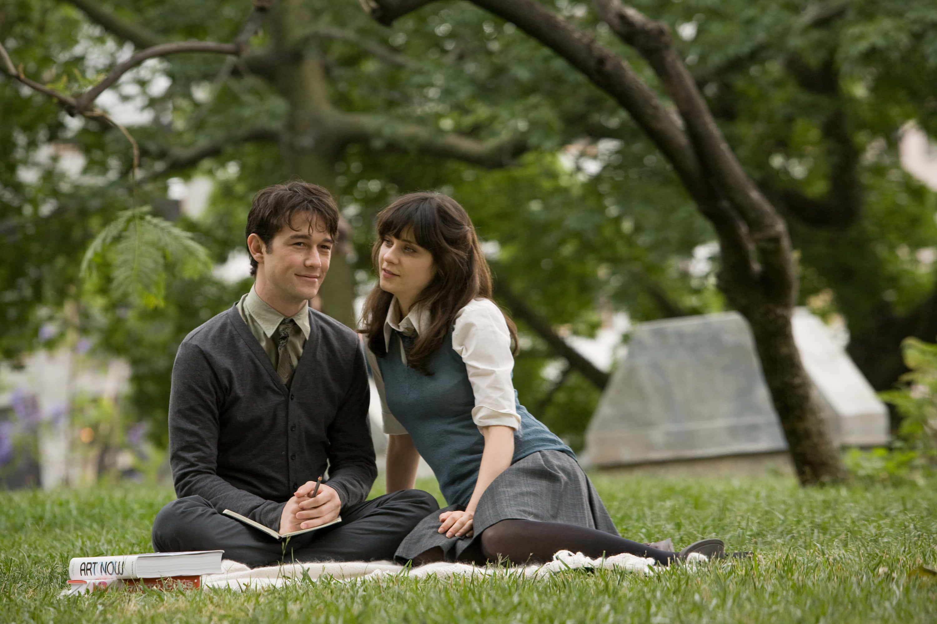500 Days Of Summer 桌布 壁紙 — 一對年輕情侶坐在陽光普照的公園裡的毯子上，周圍是茂密的綠樹和柔軟的草地。