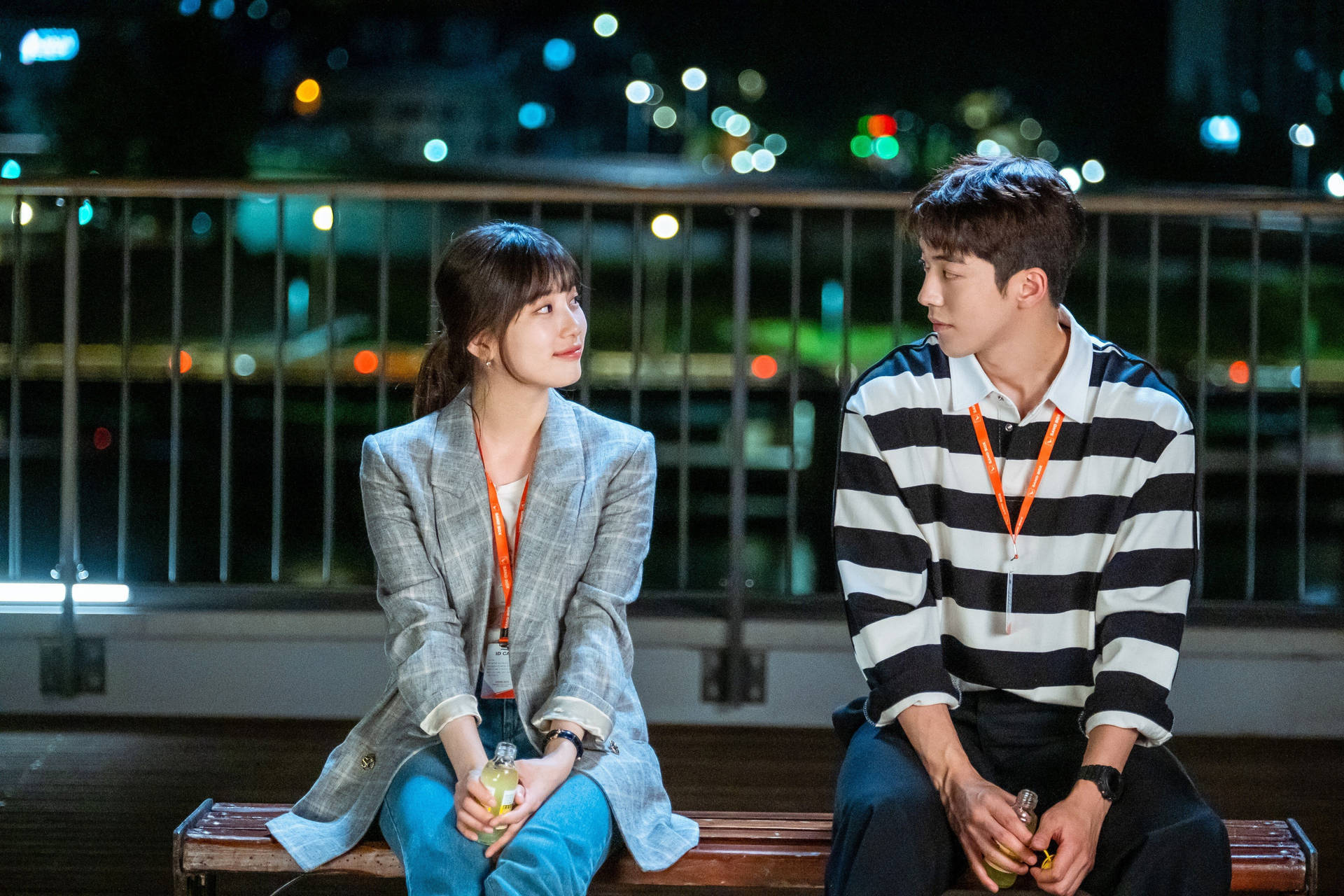 kdrama 桌布 壁紙 — 一對年輕情侶夜晚坐在長椅上，互相凝視對方的眼睛，背後的城市燈光柔和閃爍。