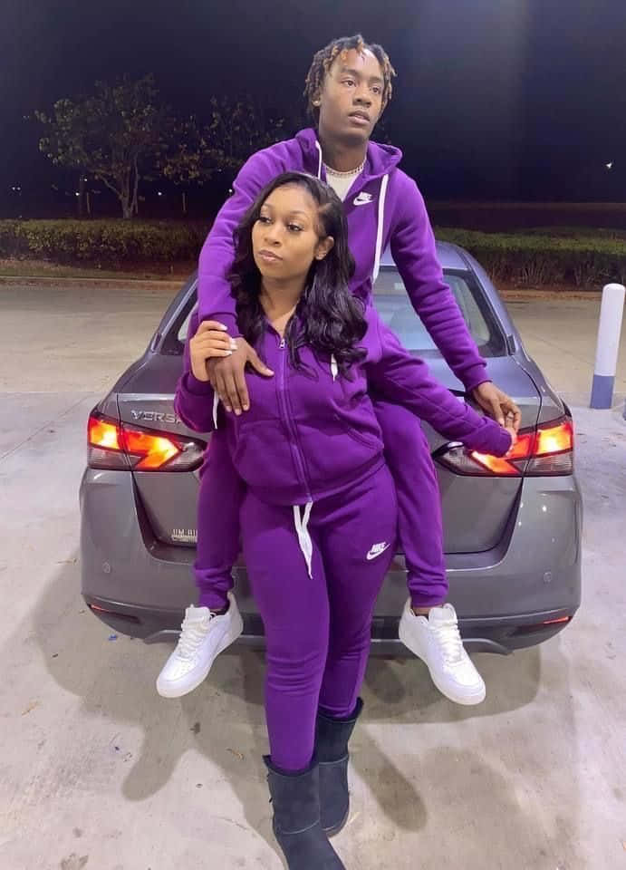 Matching Couple Profile 桌布 壁紙 — 一對年輕情侶穿著相配的紫色Nike運動套裝，在夜晚的灰色汽車前，男子騎在女子背上，俏皮地擺姿勢。