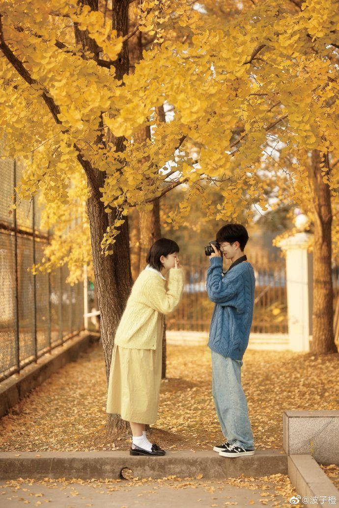 Couples 桌布 壁紙 — 一對年輕情侶站在金色秋樹下，一人拍照，另一人害羞地微笑，周圍落葉紛飛。