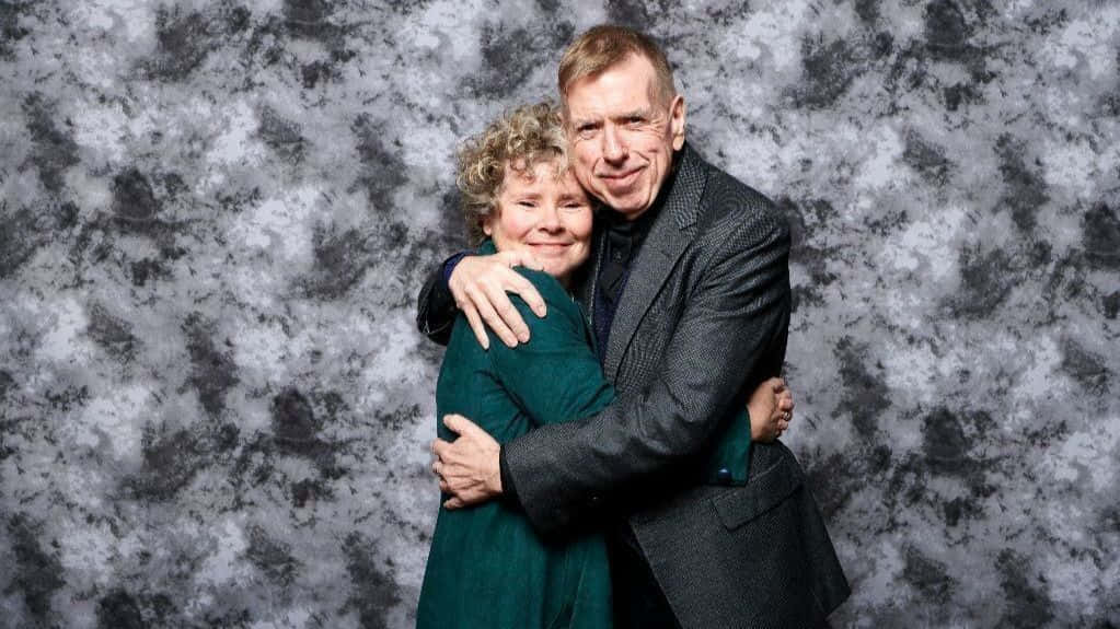 Timothy Spall 桌布 壁紙 — 一對年長夫婦在斑駁的灰白工作室背景前溫暖地擁抱，兩人都對著鏡頭溫柔微笑。