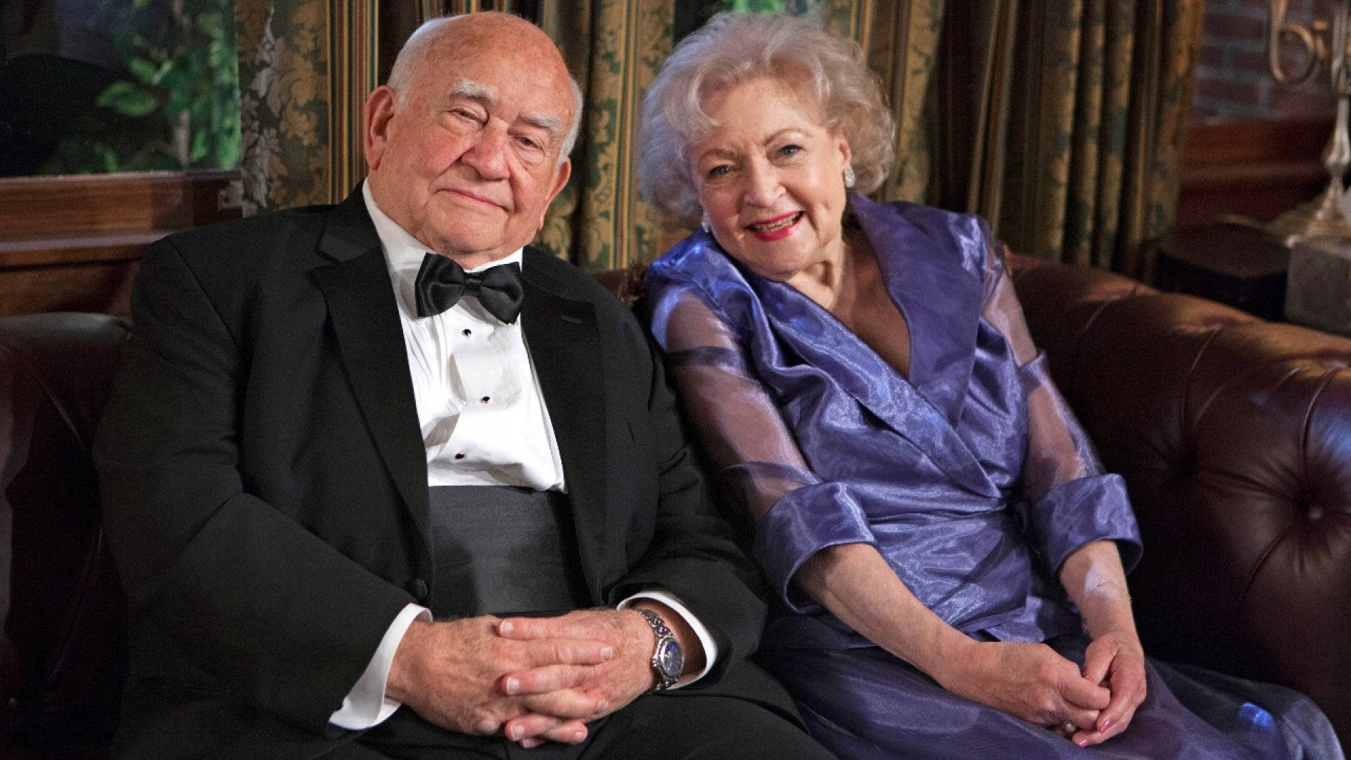 Betty White 桌布 壁紙 — 一對年長夫婦穿著正式服裝，一起坐在裝飾華麗、帶有圖案窗簾的房間裡的皮質沙發上。