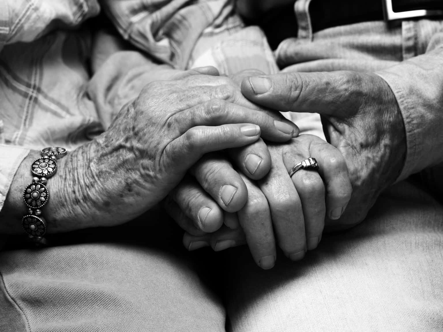 Couple Holding Hand 桌布 壁紙 — 一對年長的手溫柔地緊握在一起，戴著結婚戒指和珠串手鍊的黑白特寫。