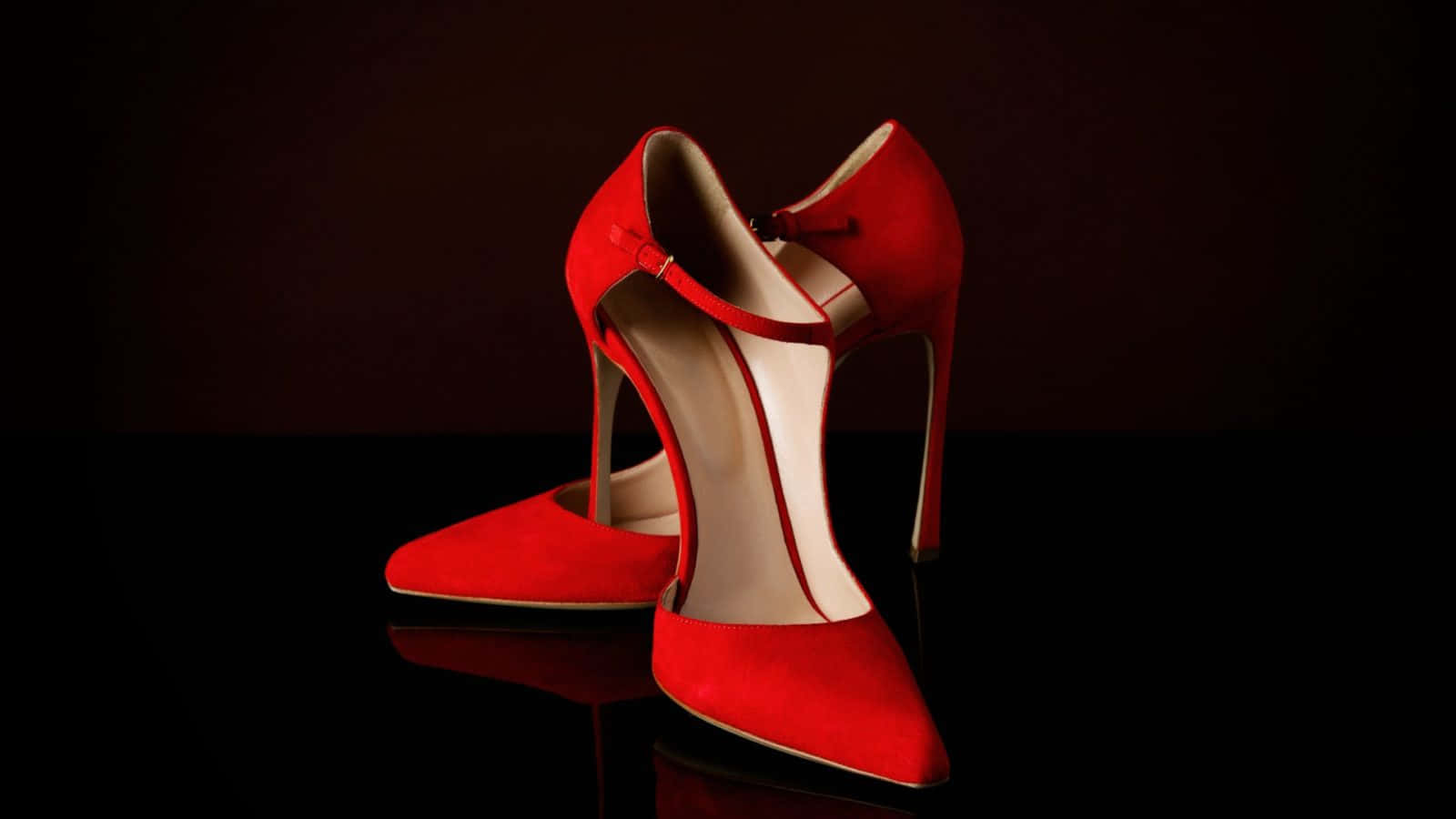 Red High Heels 桌布 壁紙 — 一對引人注目的紅色麂皮高跟鞋，配有細帶和細高跟，優雅地放置在深色背景下的光面黑色表面上。