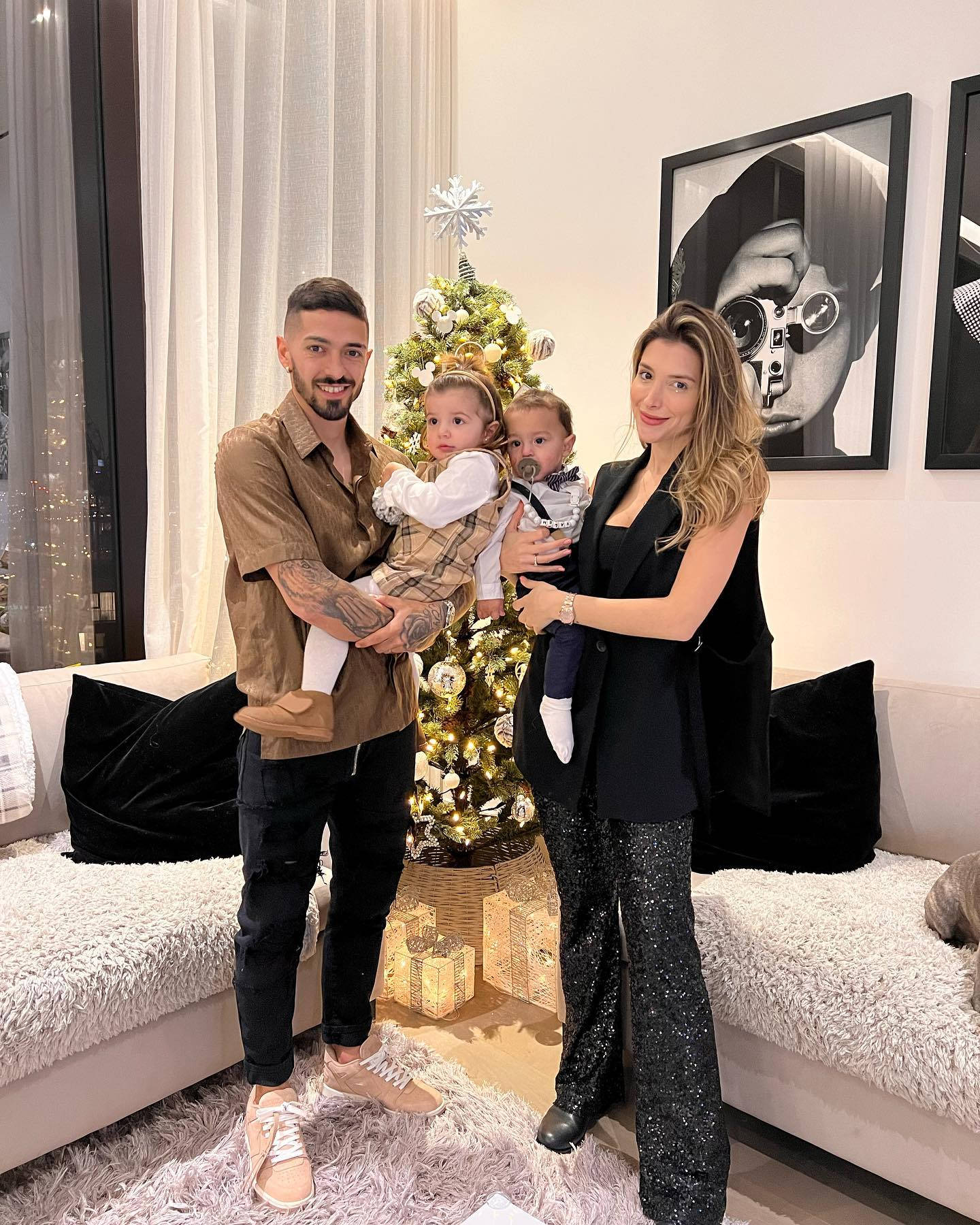 Family 桌布 壁紙 — 一對微笑夫婦與他們的兩個幼兒在裝飾好的聖誕樹旁，於舒適的現代客廳中擺姿勢。