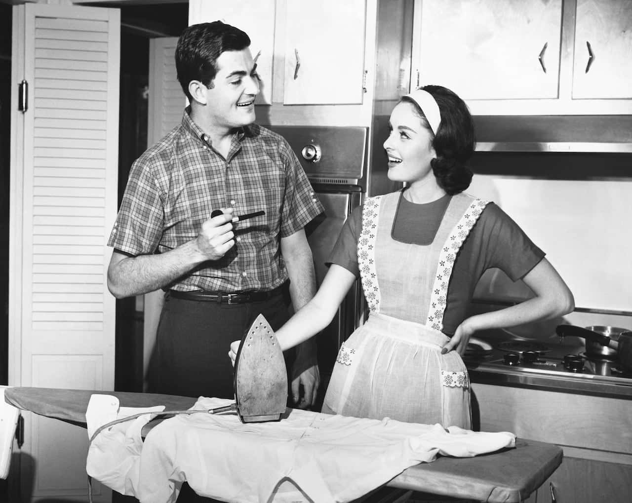 1950s 桌布 壁紙 — 一對微笑的1950年代夫婦在廚房裡，女人在熨衣服，男人拿著煙斗。