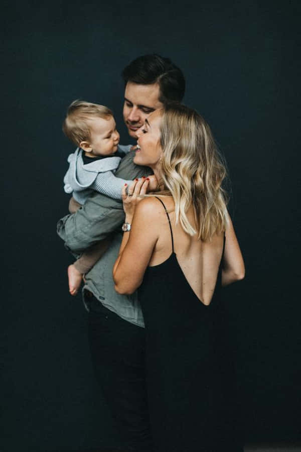 Family 桌布 壁紙 — 一對微笑的夫婦在昏暗的攝影棚背景下，用柔和的光線擁抱他們的嬰兒。