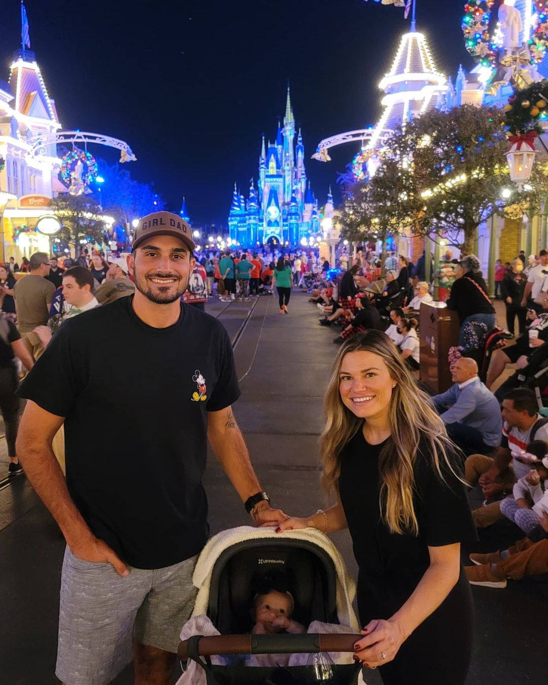Disneyland Castle 桌布 壁紙 — 一對微笑的夫婦帶著嬰兒在嬰兒車裡，晚上在燈火通明的迪士尼城堡前，周圍是節日的人群。