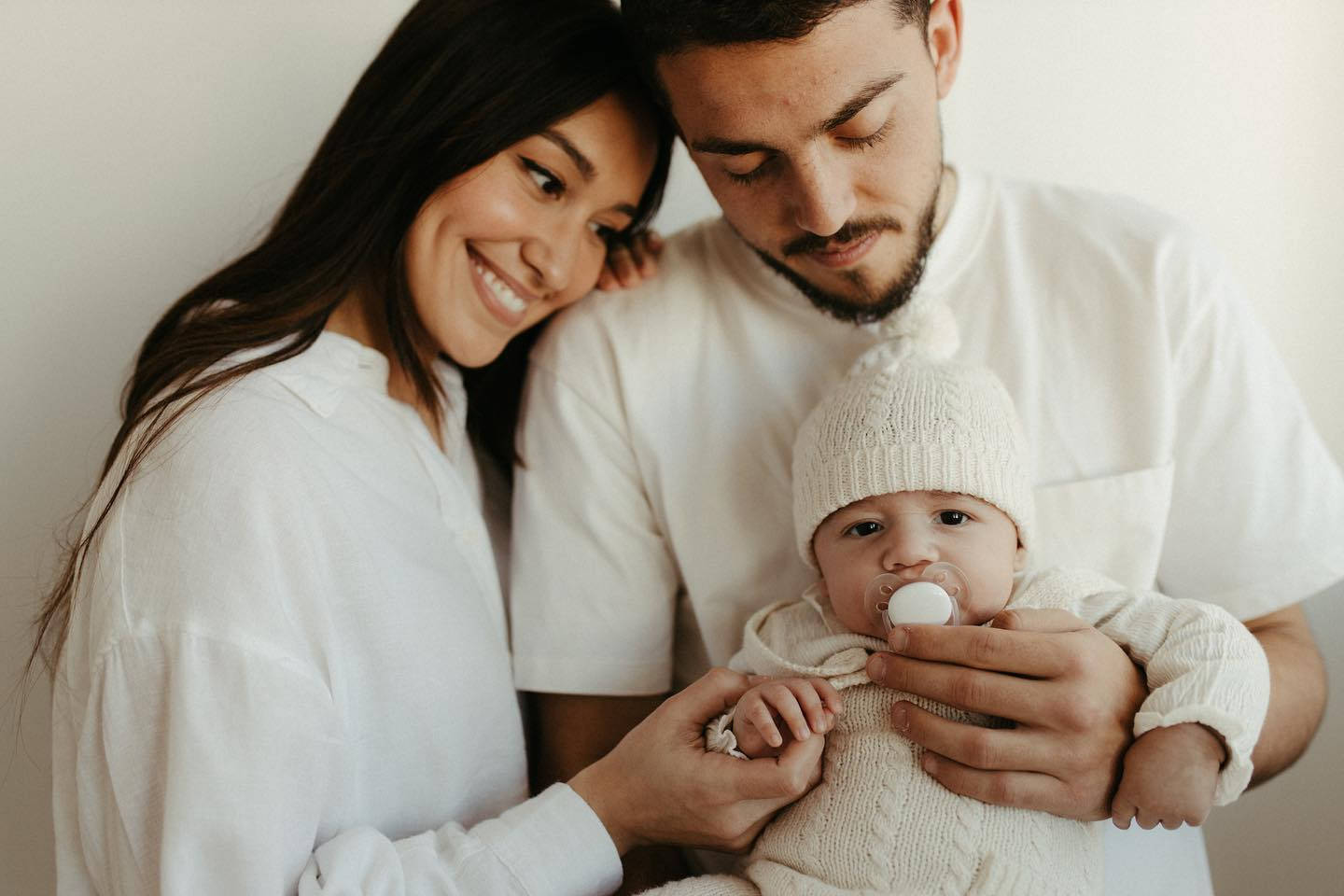 Family 桌布 壁紙 — 一對微笑的夫婦溫柔地抱著他們穿著柔軟白色針織衫的嬰兒，背景是純白牆壁。