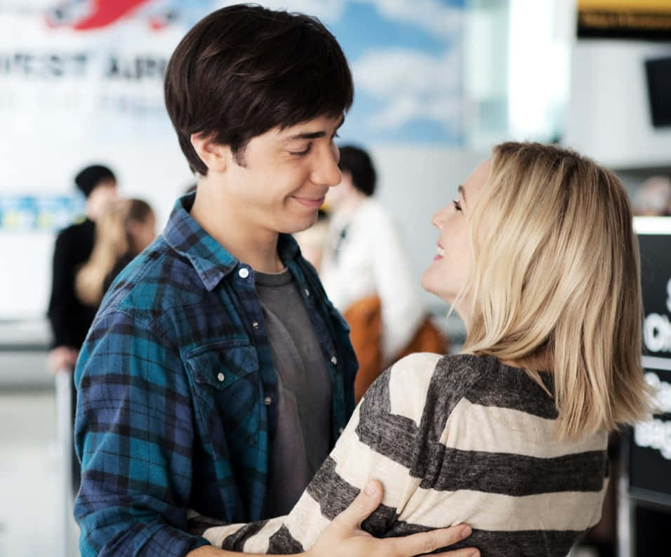 Justin Long 桌布 壁紙 — 一對微笑的年輕情侶在熙來攘往的機場航廈中熱情擁抱，背景有旅客和標示牌。