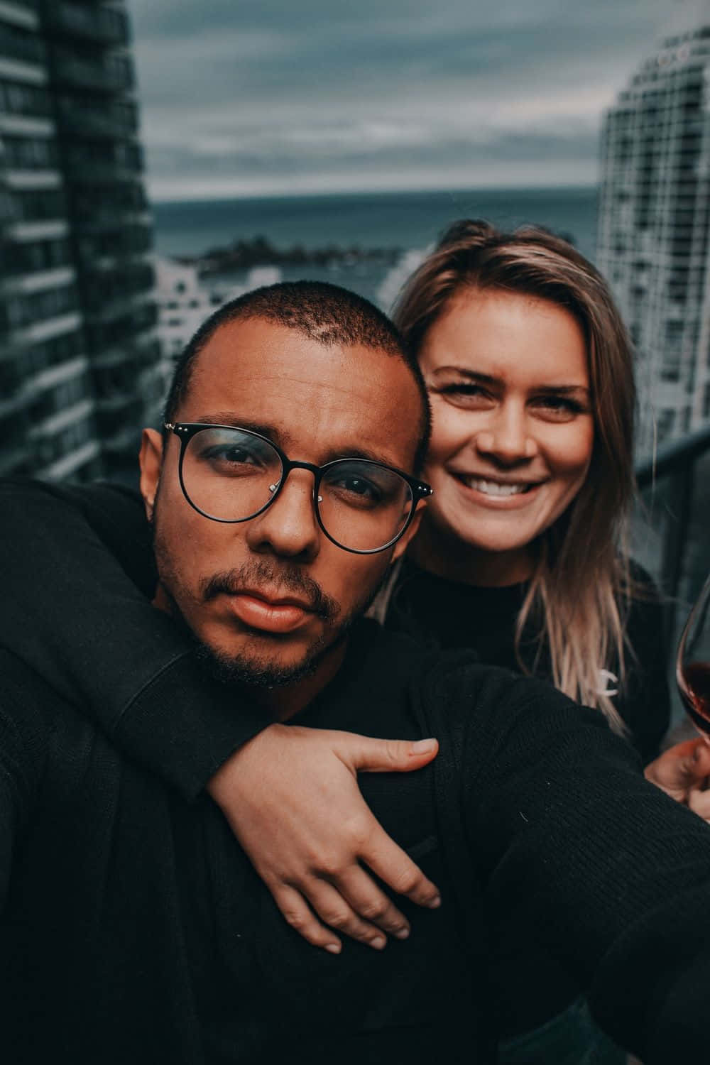 Interracial 桌布 壁紙 — 一對微笑的情侶在一個俯瞰海洋、天空多雲的城市陽台上擁抱。