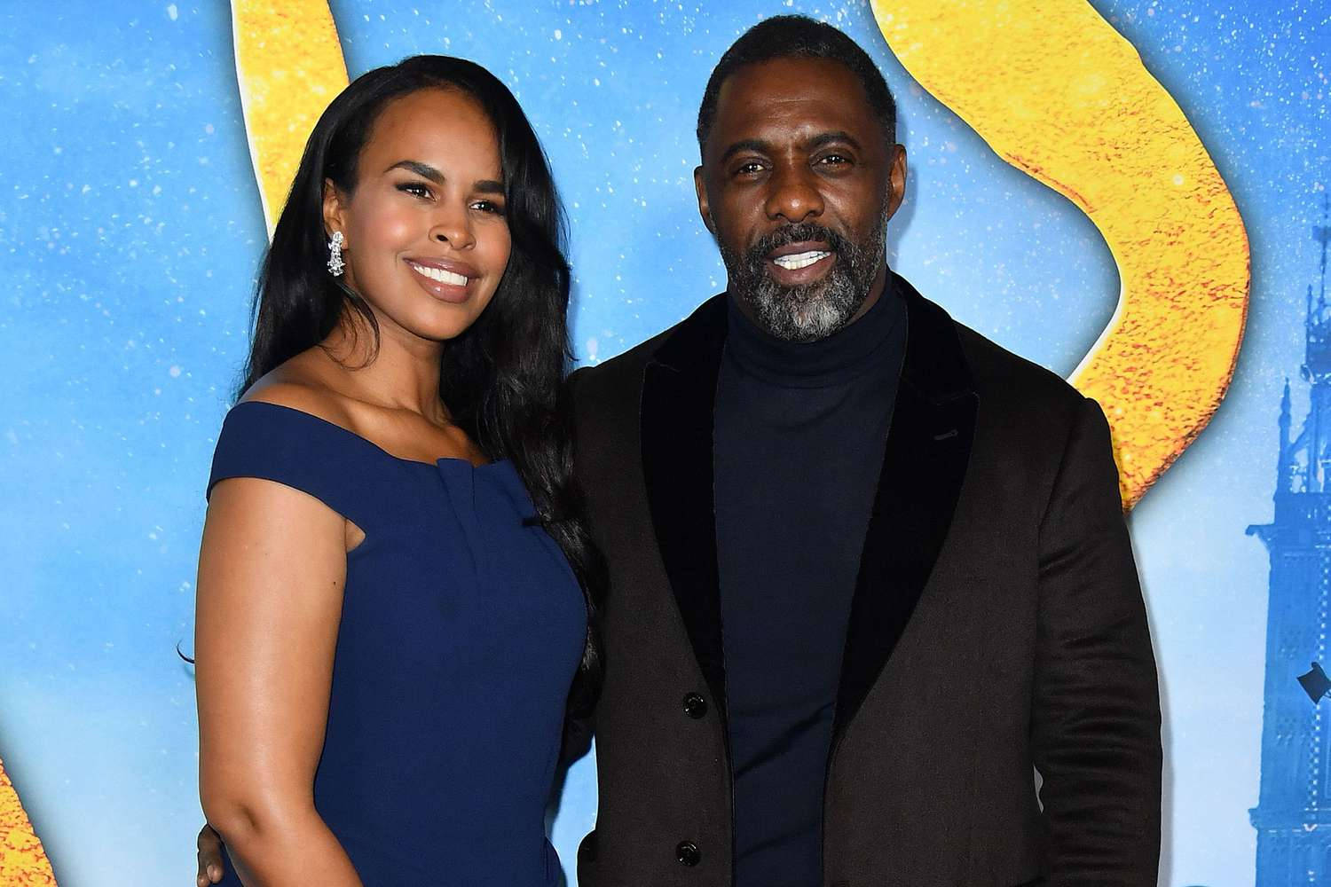 idris elba 桌布 壁紙 — 一對微笑的情侶在充滿活力的藍色和黃色電影首映背景前合影，背景有城堡輪廓。
