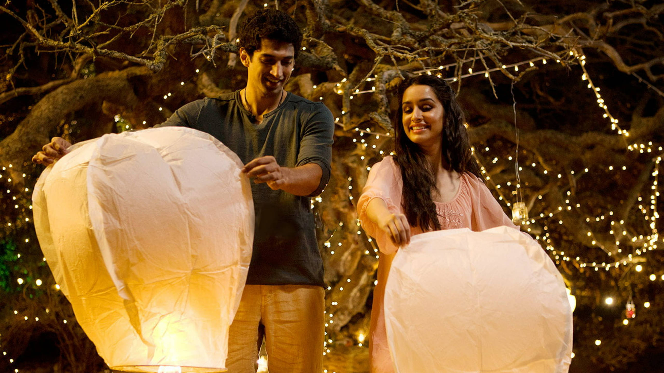 Shraddha Kapoor 桌布 壁紙 — 一對微笑的情侶在夜晚拿著發光的天燈，站在一棵裝飾著閃爍仙女燈的樹下。