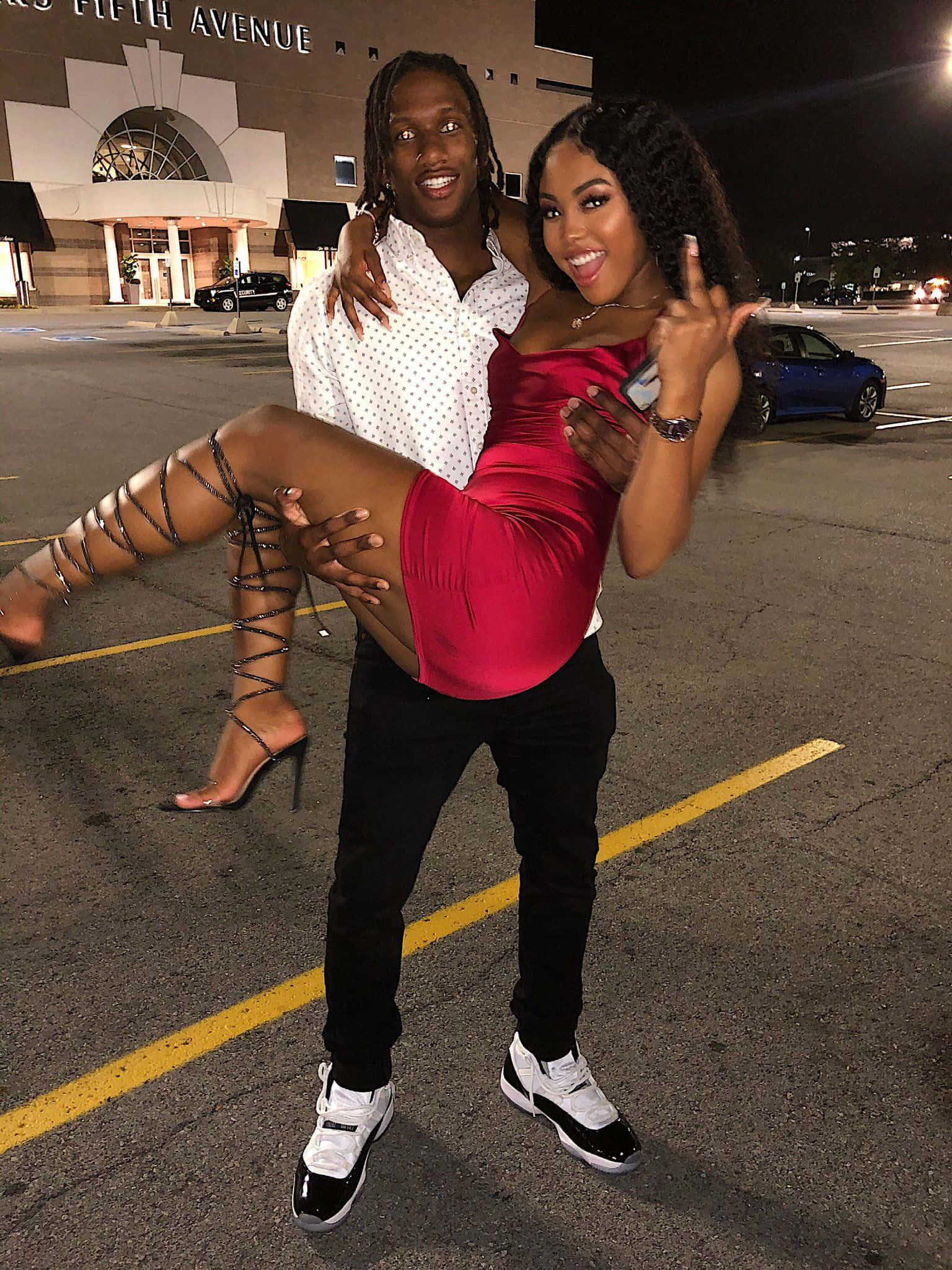 Cute Black Couple 桌布 壁紙 — 一對微笑的情侶在夜晚的停車場中俏皮地擺姿勢，背後是城市燈光和第五大道的建築。