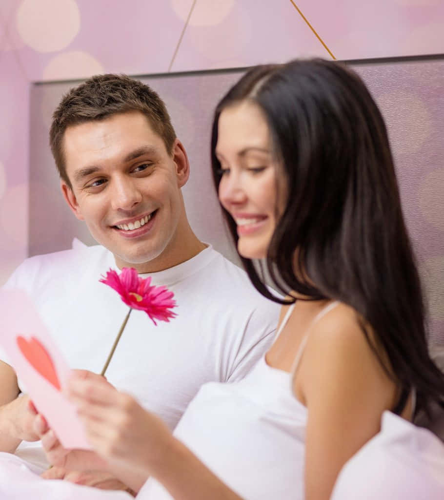 Cute Love 桌布 壁紙 — 一對微笑的情侶在床上分享一朵粉紅色的花和一張卡片，在柔和燈光和粉紅色散景燈光的浪漫臥室中。