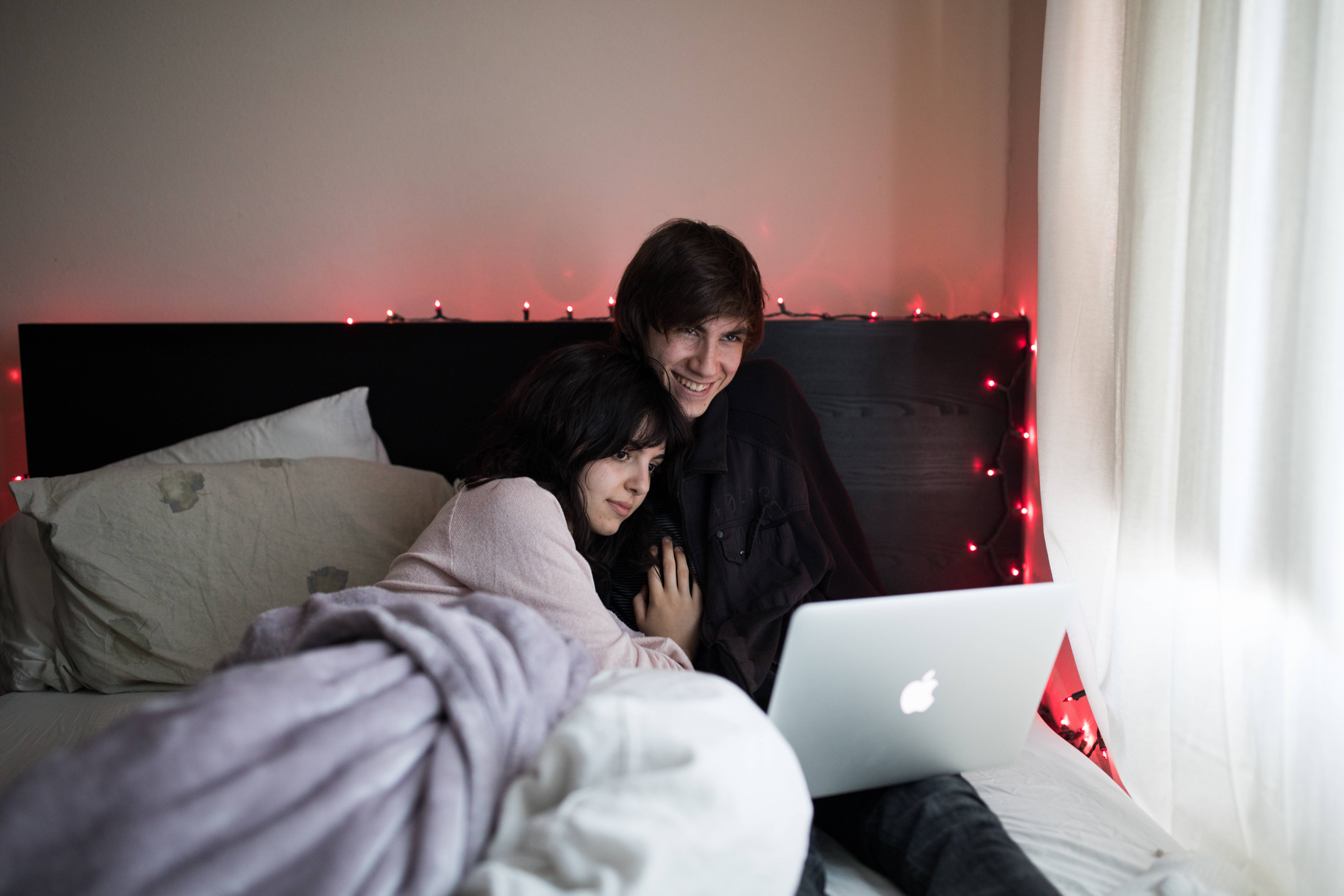 Netflix 桌布 壁紙 — 一對微笑的情侶在床上蓋著毯子依偎，看著身後有紅色串燈發光的筆記型電腦。