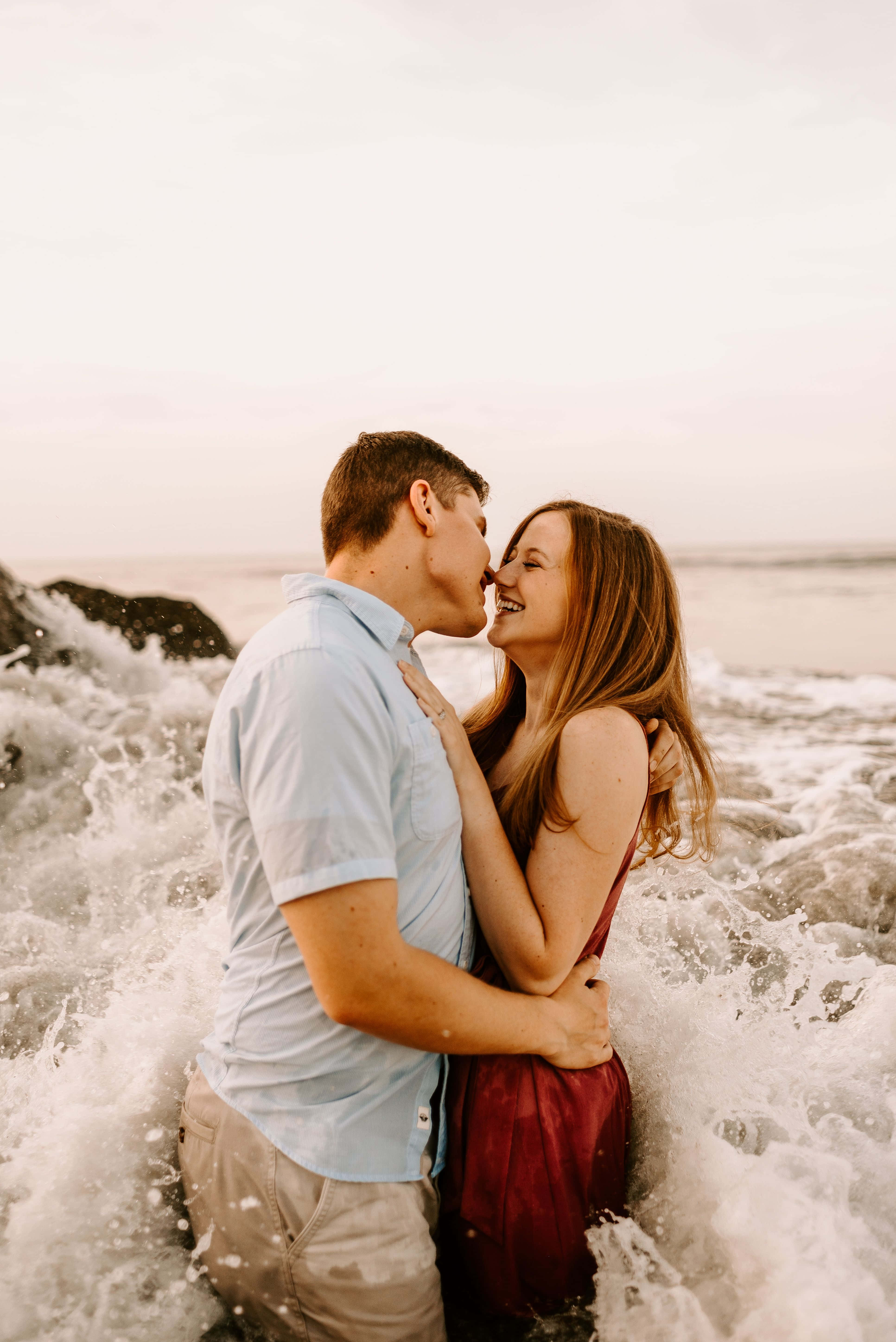 Kissing 桌布 壁紙 — 一對微笑的情侶在日落時分的岩石海岸上熱情接吻，洶湧的海浪在他們周圍飛濺。