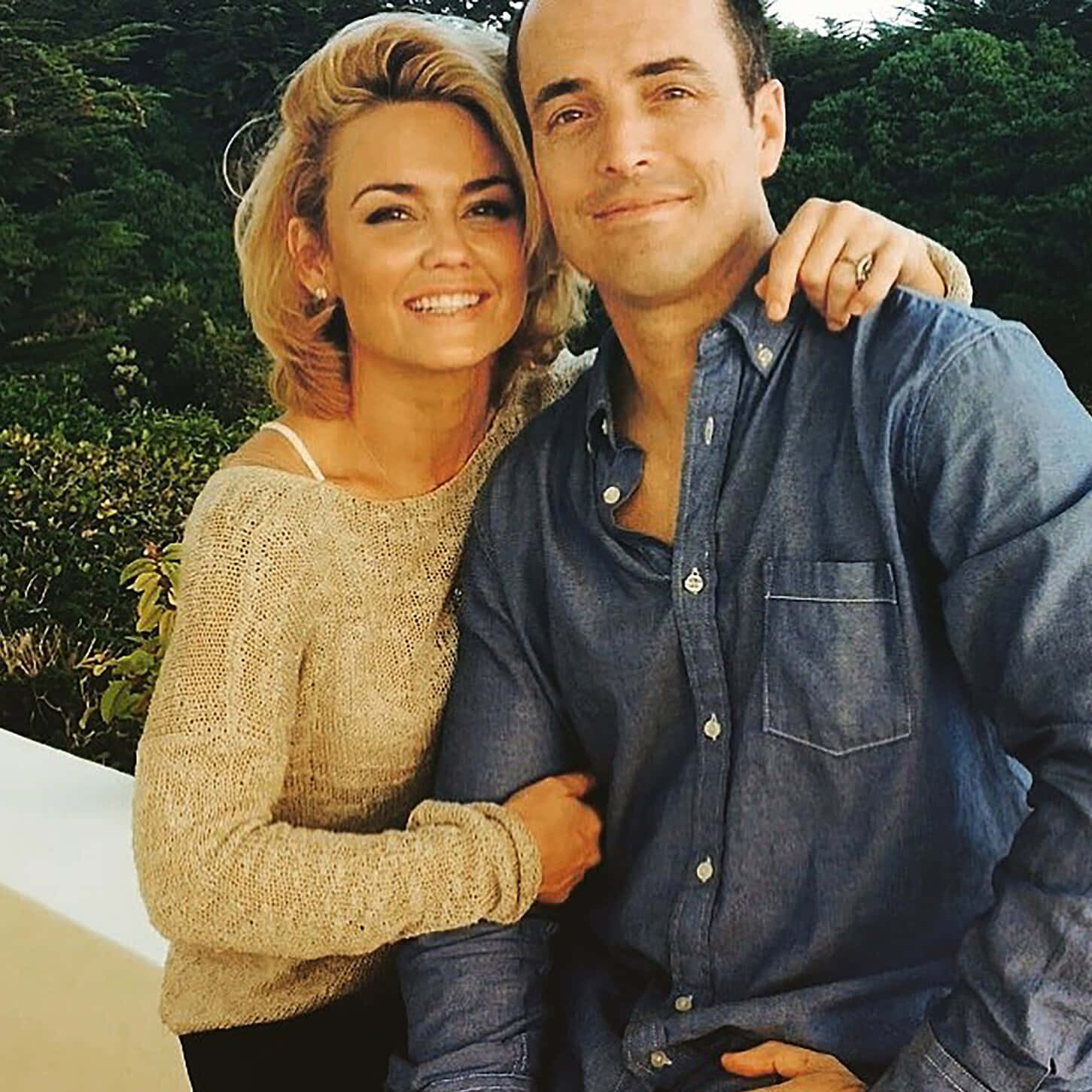 Kelly Carlson 桌布 壁紙 — 一對微笑的情侶在明亮的日光下，身後有茂密的綠植，溫暖地擁抱在一起。