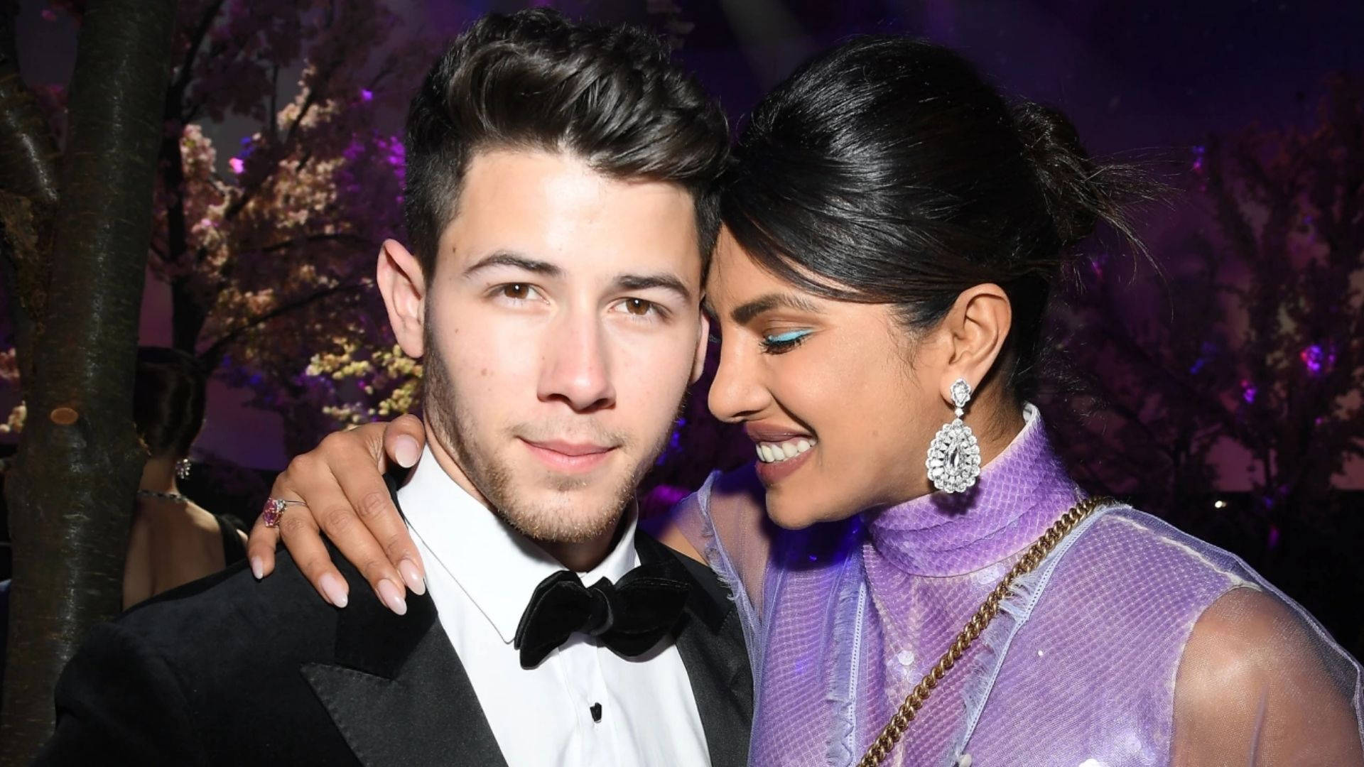 Nick Jonas 桌布 壁紙 — 一對微笑的情侶在晚間活動中緊密地擺姿勢，背景有紫色燈光和盛開的樹木。