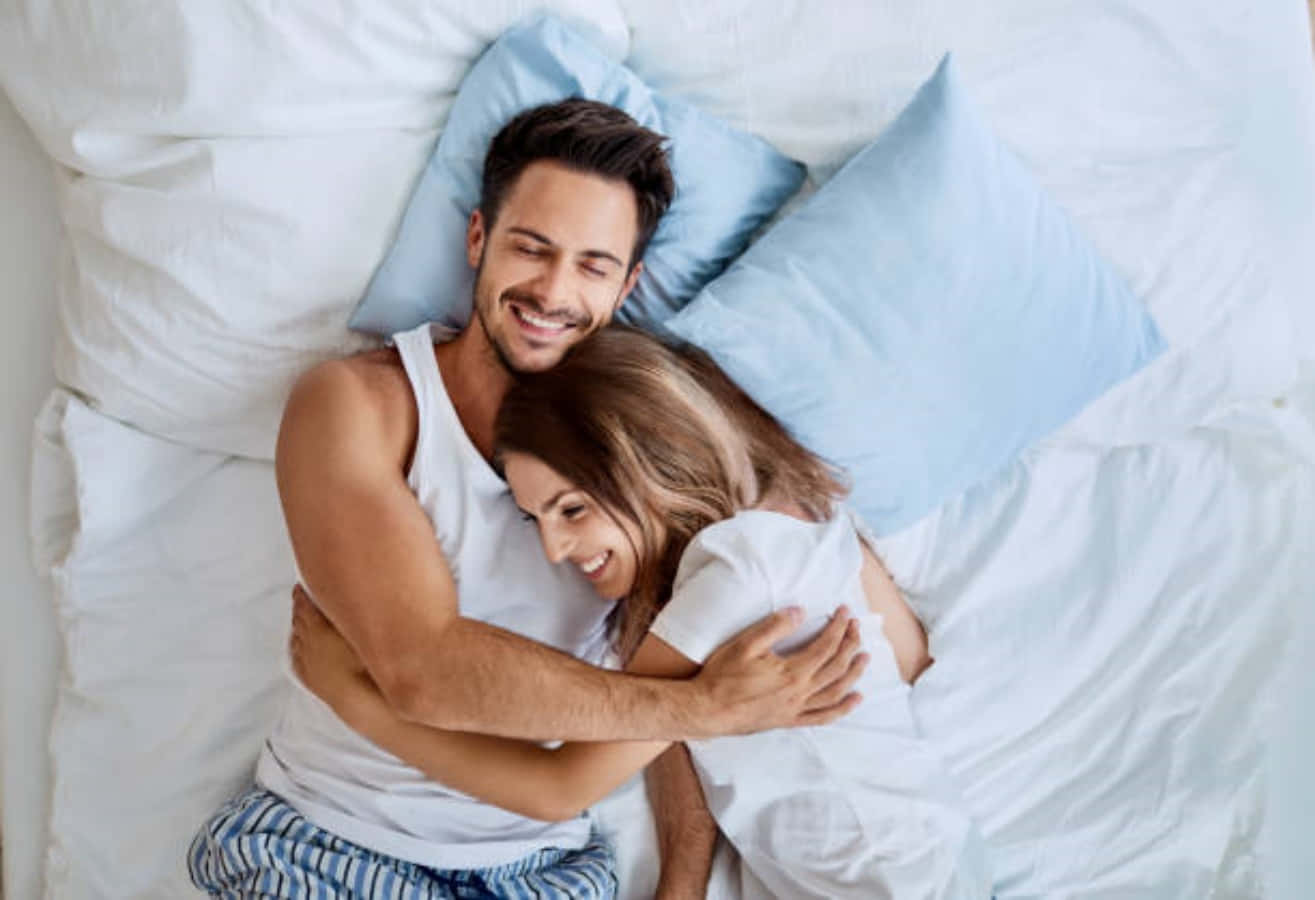Cuddle 桌布 壁紙 — 一對微笑的情侶在柔軟的白色床單和淺藍色枕頭下，在床上溫暖地擁抱。