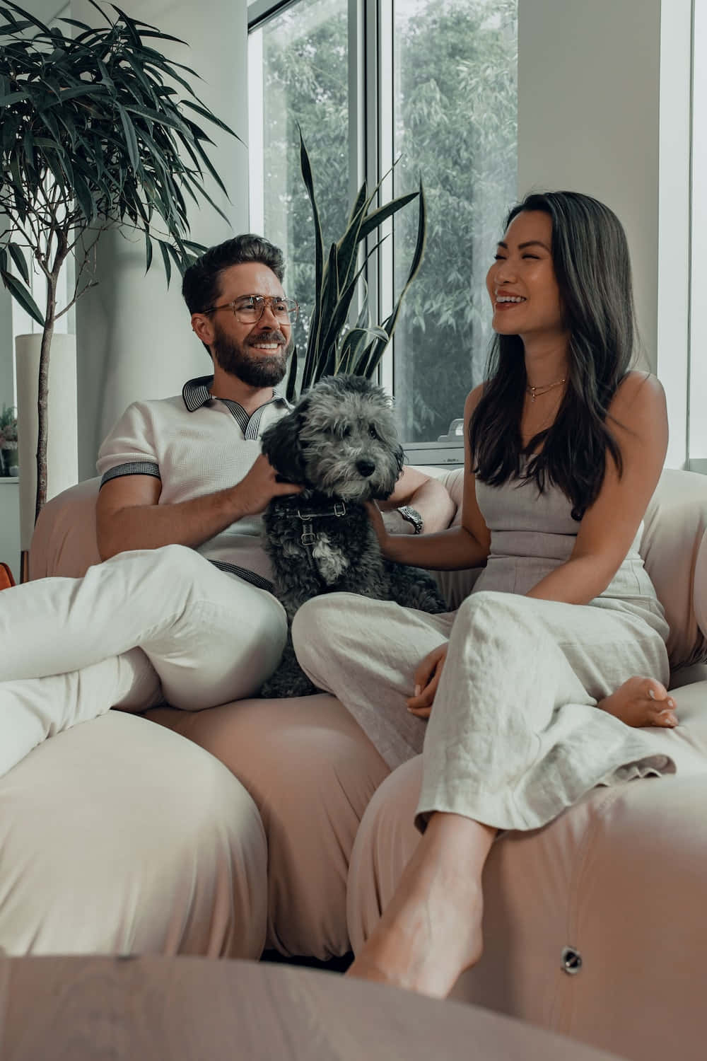 Beautiful Couple 桌布 壁紙 — 一對微笑的情侶在米色沙發上放鬆，他們之間有一隻毛茸茸的灰色狗，被大窗戶透進的柔和自然光沐浴著。