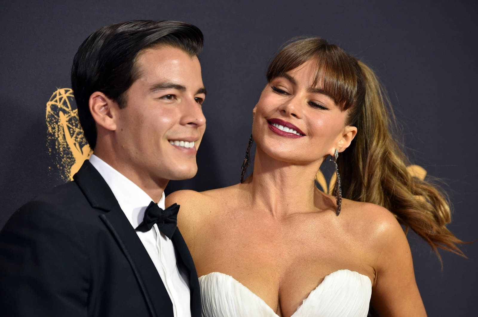 Sofia Vergara 桌布 壁紙 — 一對微笑的情侶在紅地毯上擺姿勢，背景為深色，帶有金色艾美獎標誌。