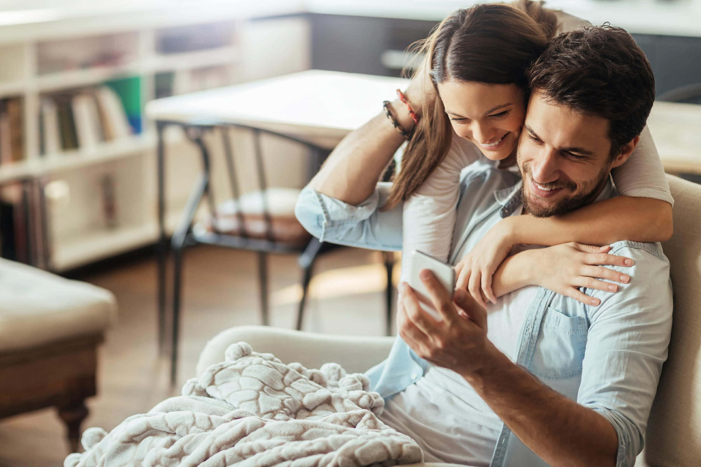 Love Couple 桌布 壁紙 — 一對微笑的情侶在舒適的客廳裡緊緊依偎，一起看著手機。