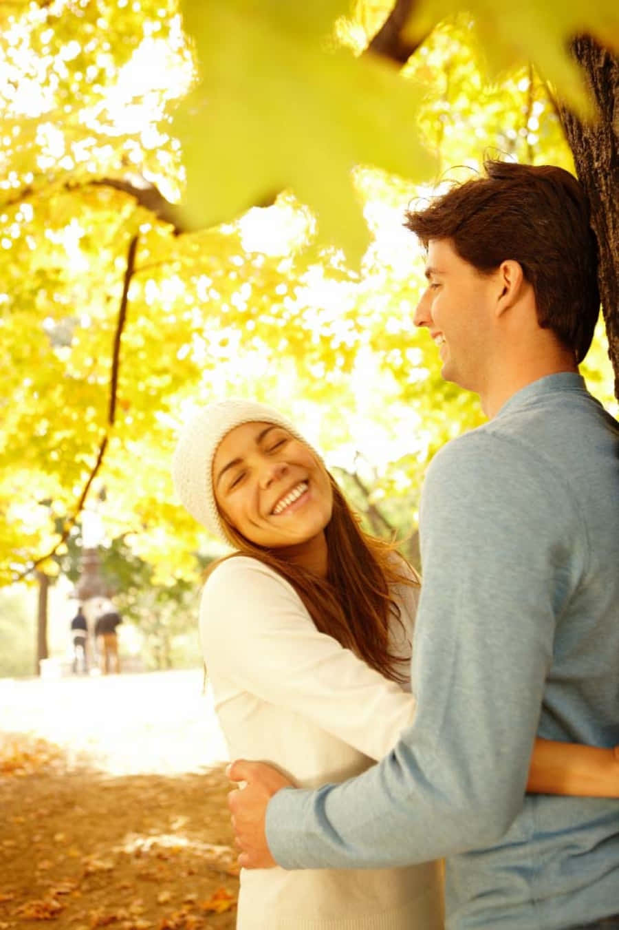 Falling In Love 桌布 壁紙 — 一對微笑的情侶在陽光明媚的公園裡，在金黃色的秋葉下擁抱，溫暖的光線穿過樹木。