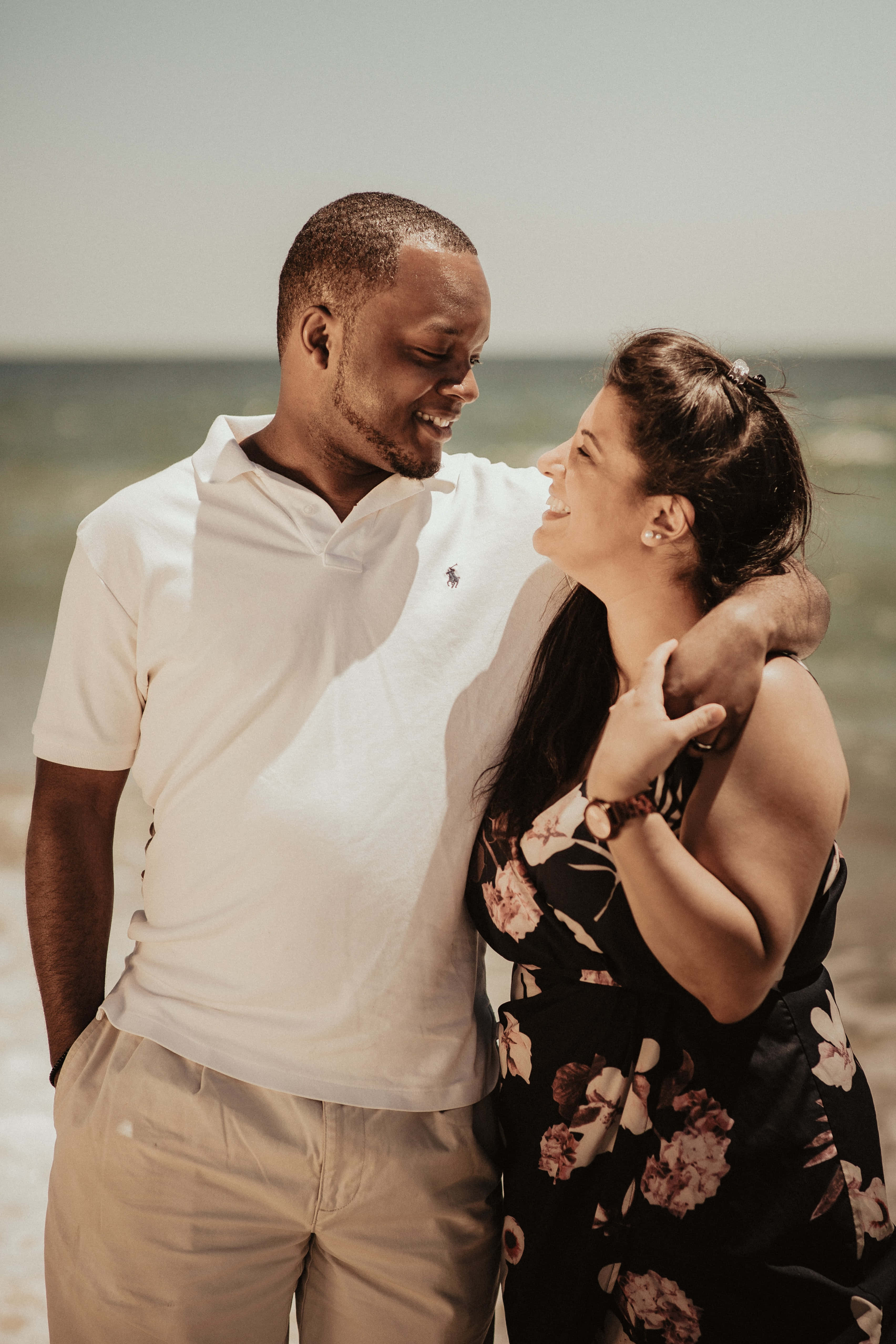 Interracial 桌布 壁紙 — 一對微笑的情侶在陽光明媚的海灘上擁抱，身後是海浪。
