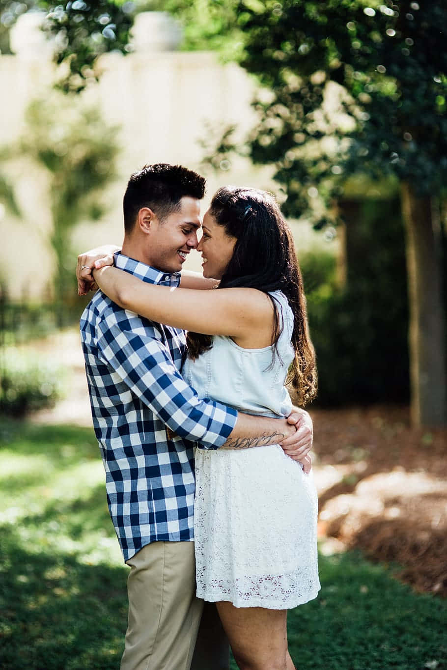couple love 桌布 壁紙 — 一對微笑的情侶在陽光明媚的花園中溫柔擁抱，身後是斑駁的綠色樹葉。