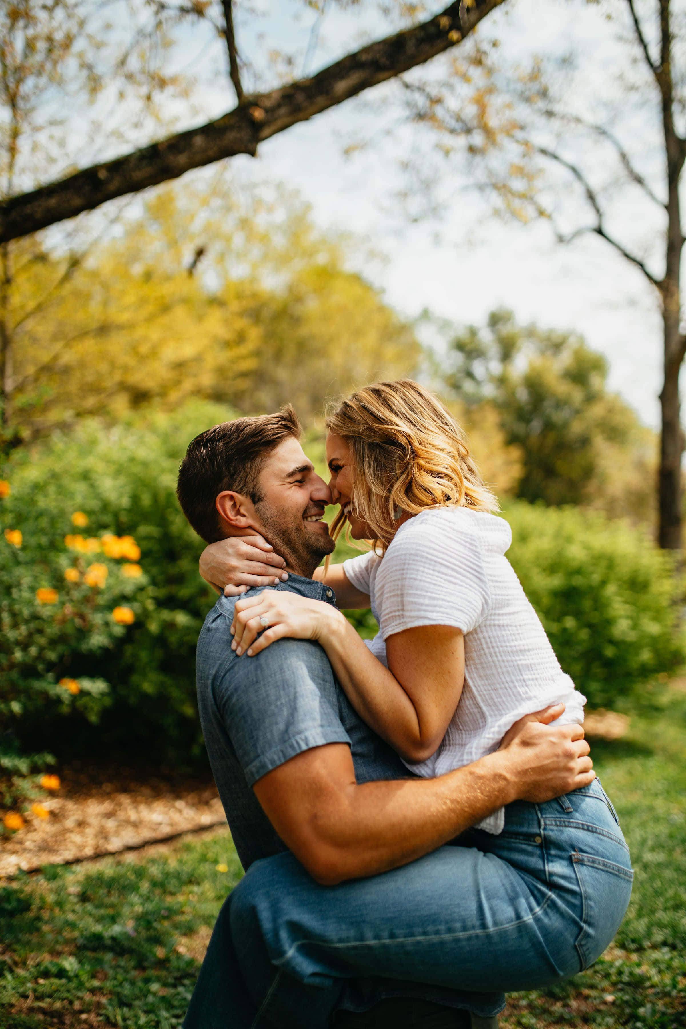 Couples 桌布 壁紙 — 一對微笑的情侶在陽光明媚的花園中溫柔擁抱，周圍有金色的秋葉和盛開的橙色花朵。