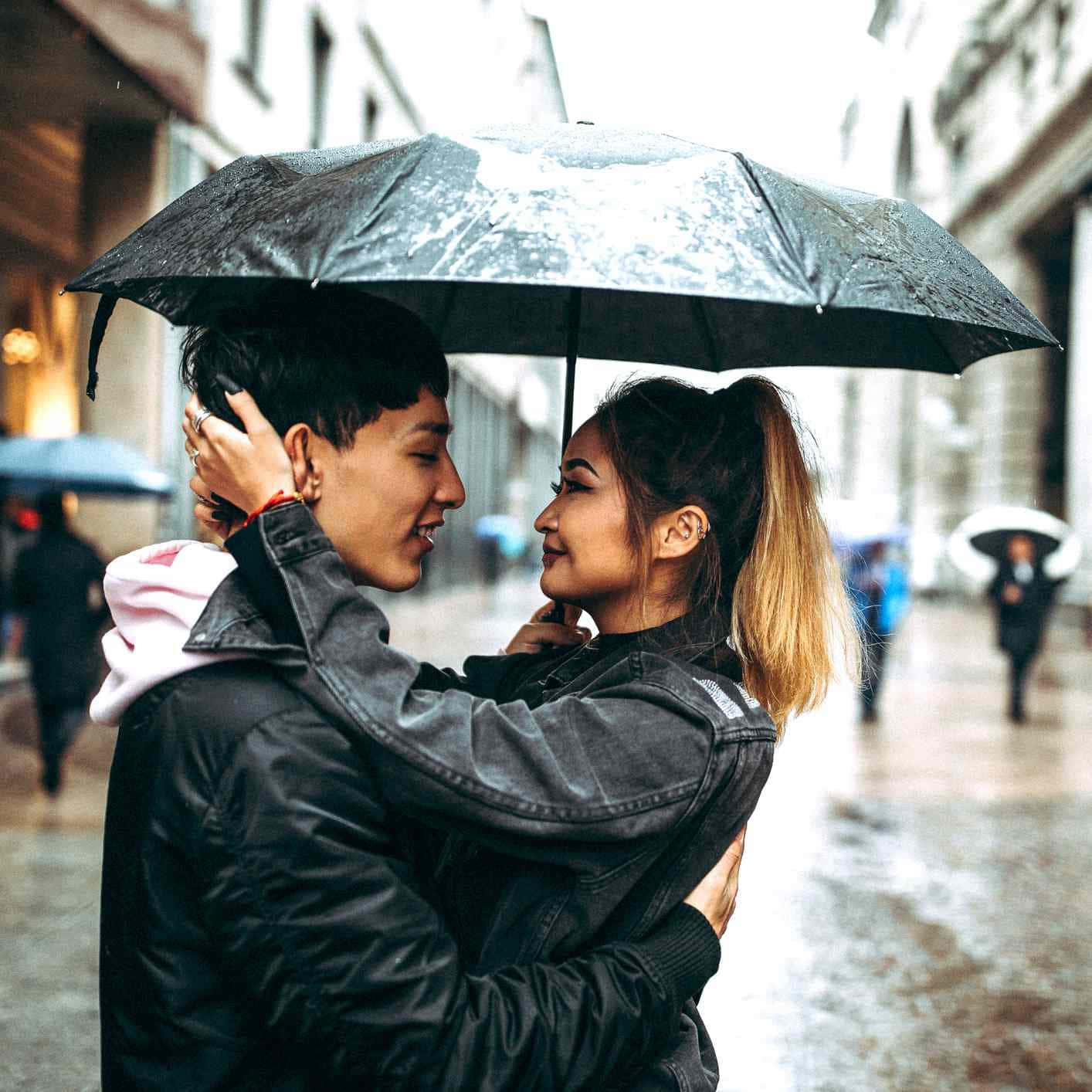 Couple In Rain 桌布 壁紙 — 一對微笑的情侶在雨天的城市街道上，在黑色雨傘下擁抱，濕漉漉的人行道和模糊的行人。